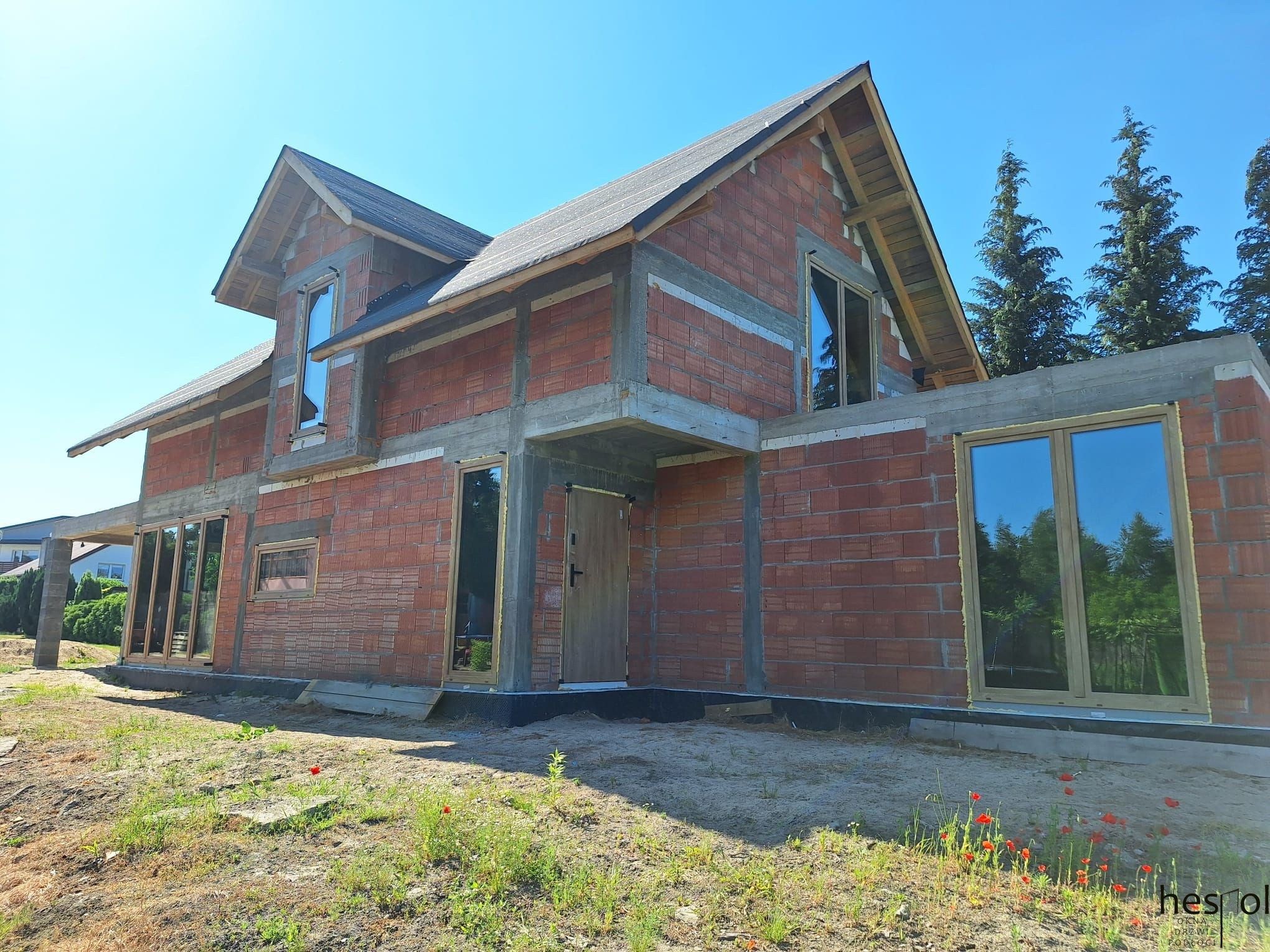 Dom w trakcie budowy z zamontowanymi oknami i drzwiami. Widoczne czerwone cegły i betonowe elementy konstrukcyjne. Na pierwszym planie trawa i kwiaty.