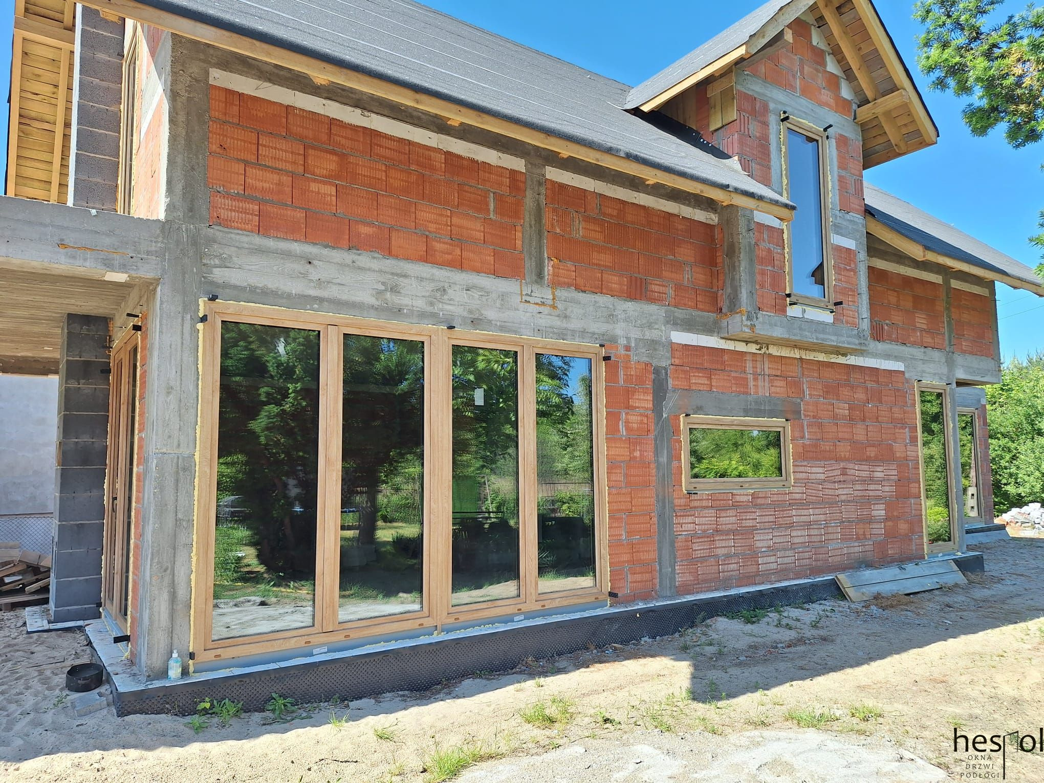 Surowy stan budynku z zamontowanymi oknami o drewnianych ramach. Widoczne cegły i betonowe elementy konstrukcyjne. Odbicie zieleni w szybach.