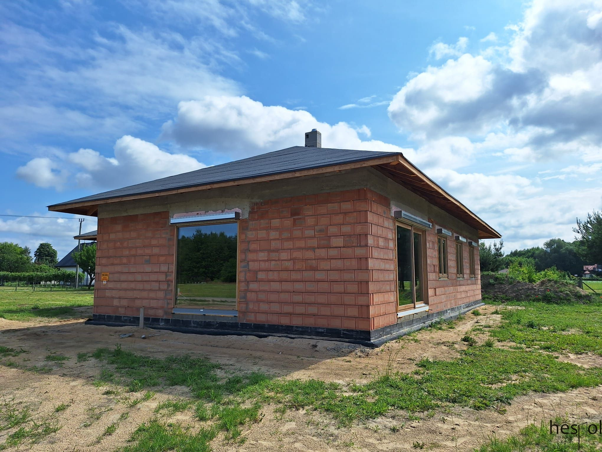 Dom w trakcie budowy, widoczne okna z drewnianą ramą, ściany z czerwonej cegły i czarny dach na tle błękitnego nieba z chmurami.