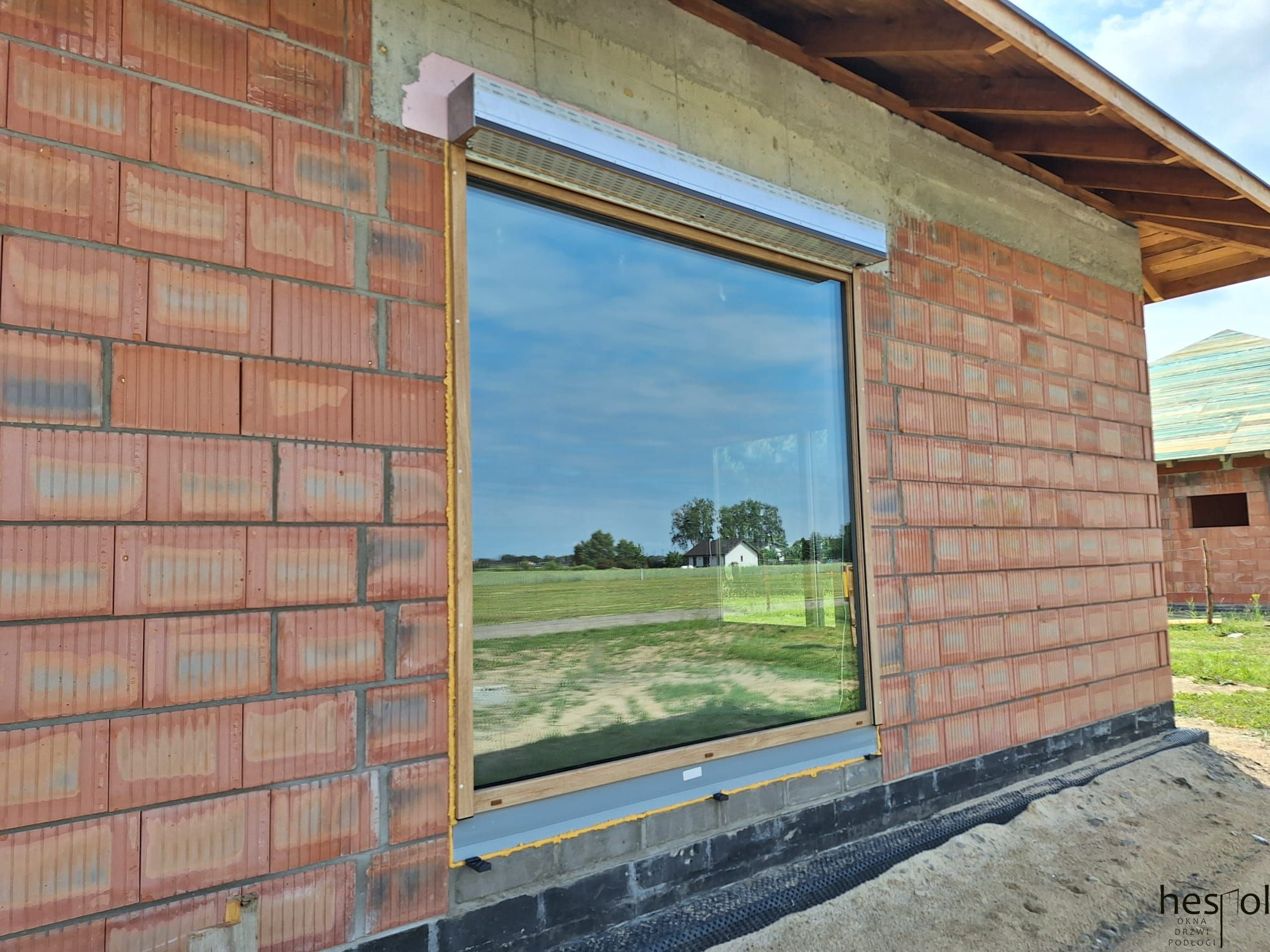 Montaż okna z roletą zewnętrzną w surowym budynku z cegły. Widoczne detale montażu, odbicie krajobrazu w szybie. Ujęcie z bliska, jasne oświetlenie.