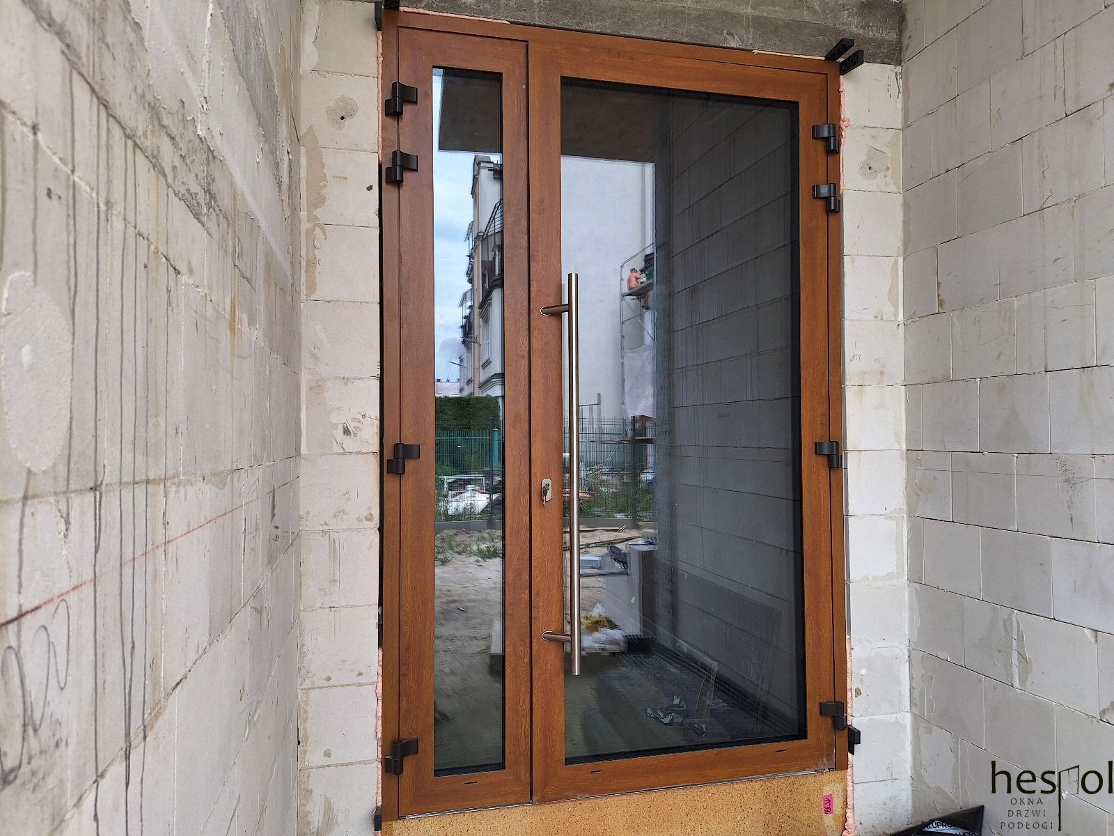 Brązowe drzwi zewnętrzne z długim, srebrnym uchwytem, zamontowane w surowym murze z betonu komórkowego. Widoczne czarne zawiasy.