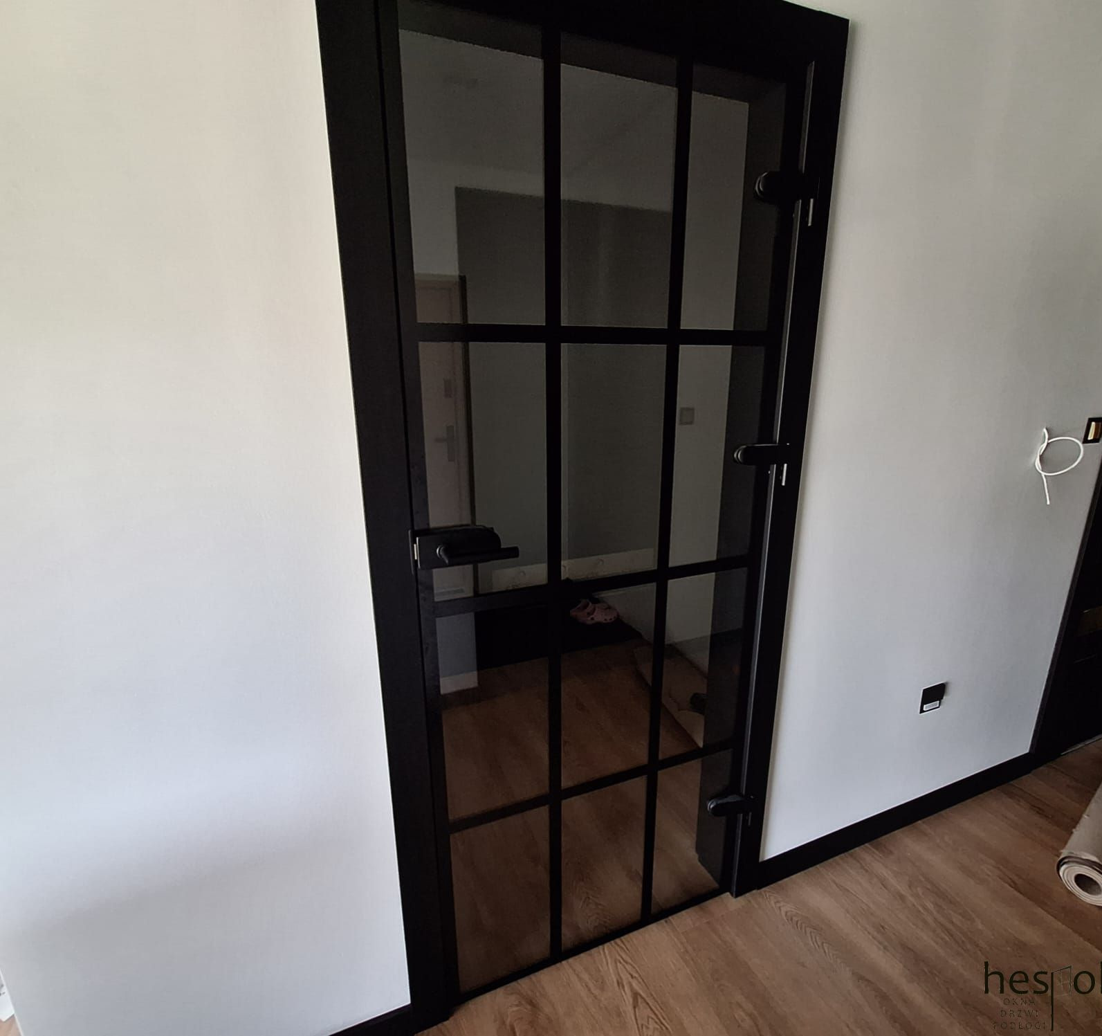 Czarne, szklane drzwi wewnętrzne z podziałami, widok na wnętrze z drewnianą podłogą, białe ściany. Minimalistyczny design z widocznymi zawiasami.
