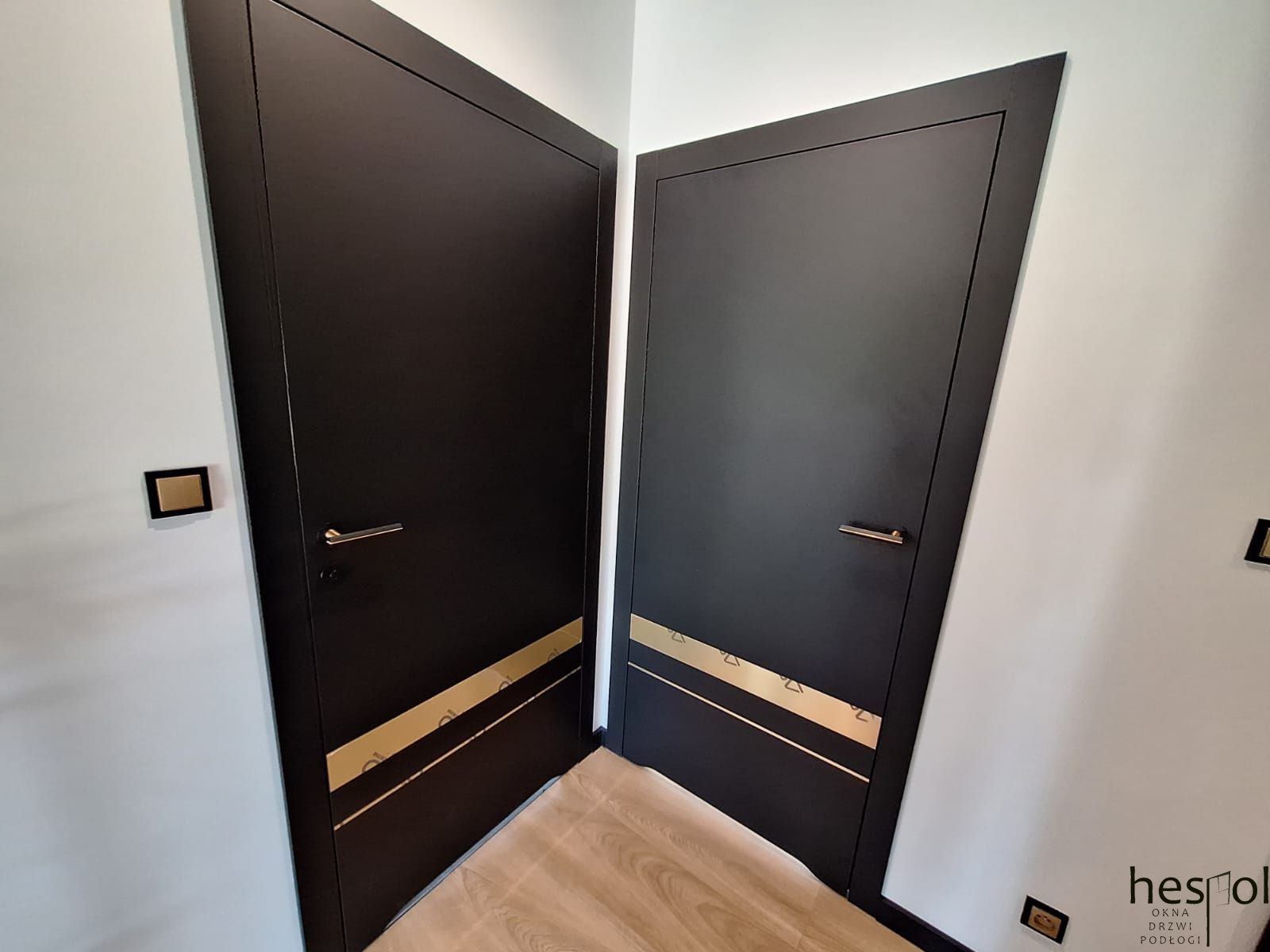 Dwa czarne, nowoczesne drzwi wewnętrzne z poziomym, złotym zdobieniem, ustawione pod kątem w rogu pomieszczenia. Widoczna klamka i kontakt. Podłoga drewniana.