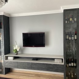 Rafał kloch - Nowoczesna szafka RTV z szarymi frontami i kamiennym blatem, flankowana przeszklonymi witrynami z kolekcją alkoholi, oświetlona kryształowym żyrandolem.