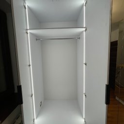 Rafał kloch - Wnętrze białej szafy wnękowej z otwartymi drzwiami, oświetleniem LED po bokach i drążkiem na ubrania.