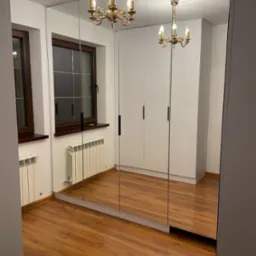 Biała szafa wnękowa z lustrzanymi drzwiami, odbijająca klasyczny żyrandol w pokoju z drewnianą podłogą i oknami z ciemnymi ramami.