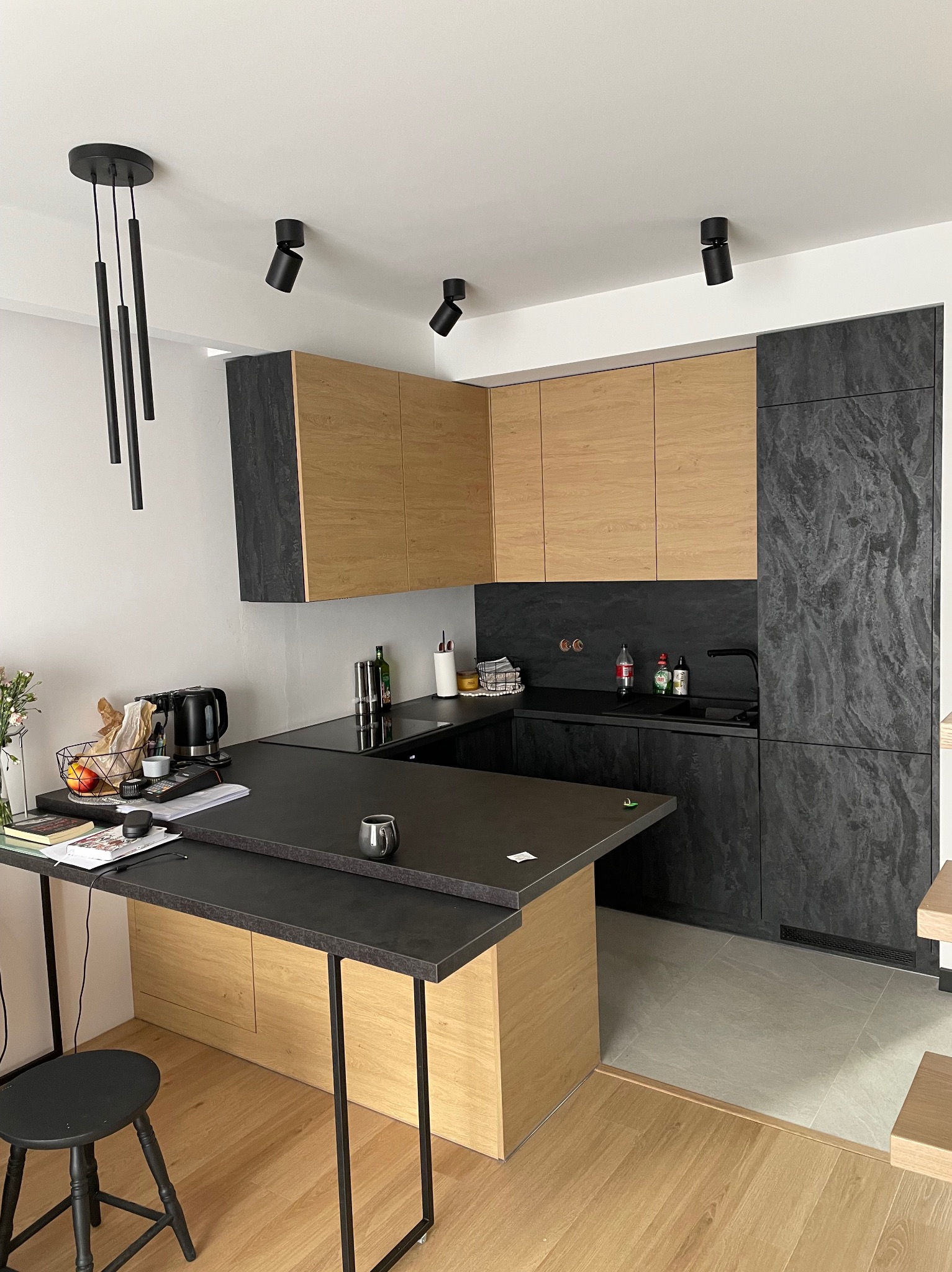 Nowoczesna kuchnia z drewnianymi i grafitowymi frontami, czarnym blatem i wiszącymi lampami, widok na wyspę kuchenną z hokerem.