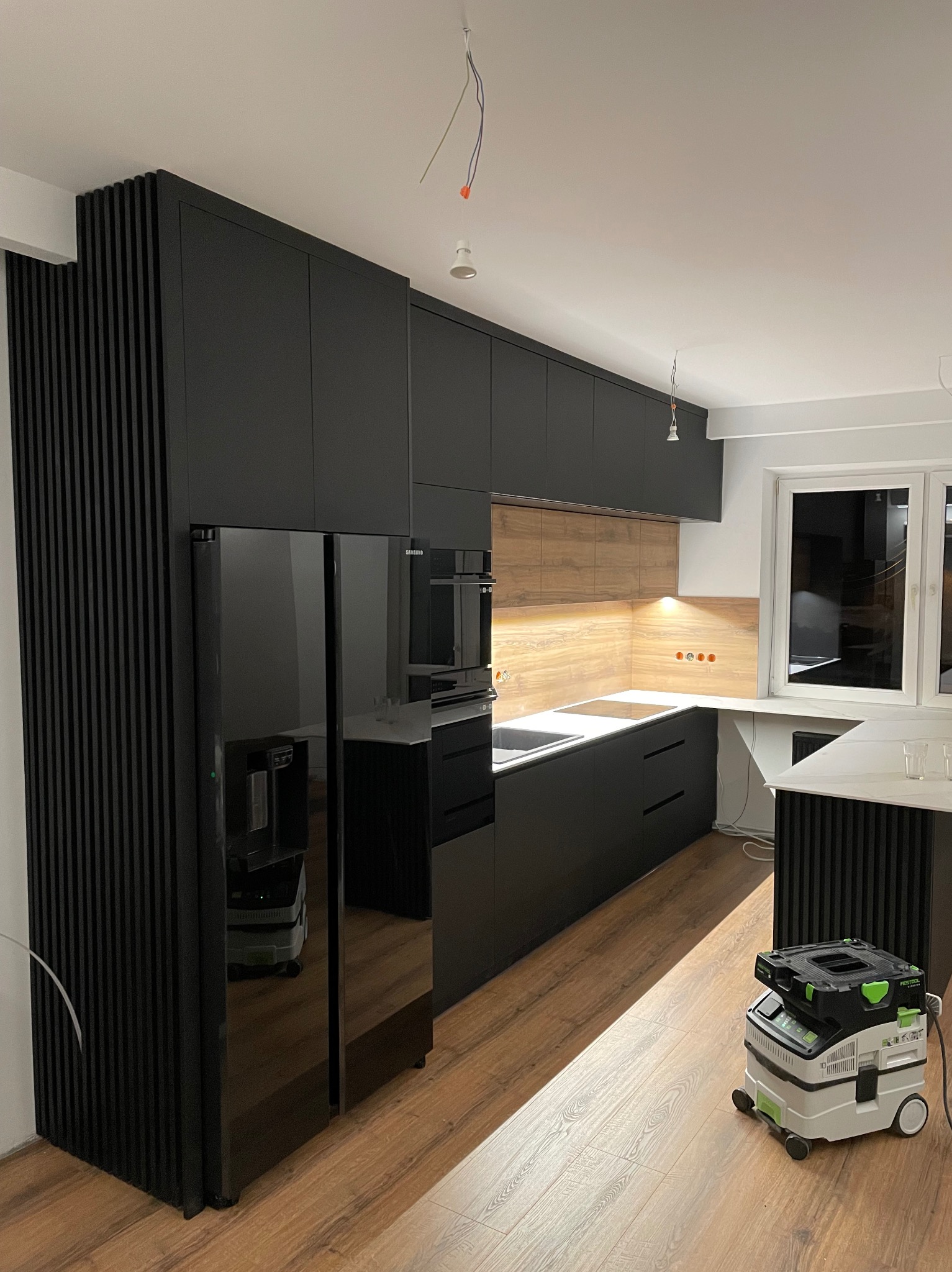 Nowoczesna kuchnia z czarnymi, lakierowanymi frontami i drewnianym panelem nad blatem, widoczny odkurzacz przemysłowy Festool, lampa wisząca bez klosza i niedokończone prace wykończeniowe.