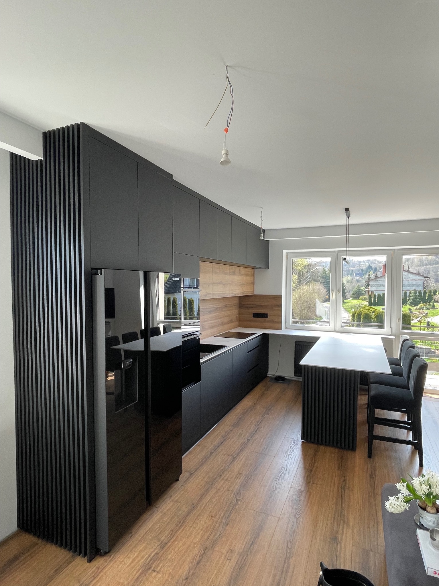 Nowoczesna kuchnia z czarnymi frontami, drewnianym panelem ściennym i lodówką w zabudowie z lamelami, widok na ogród przez okno.