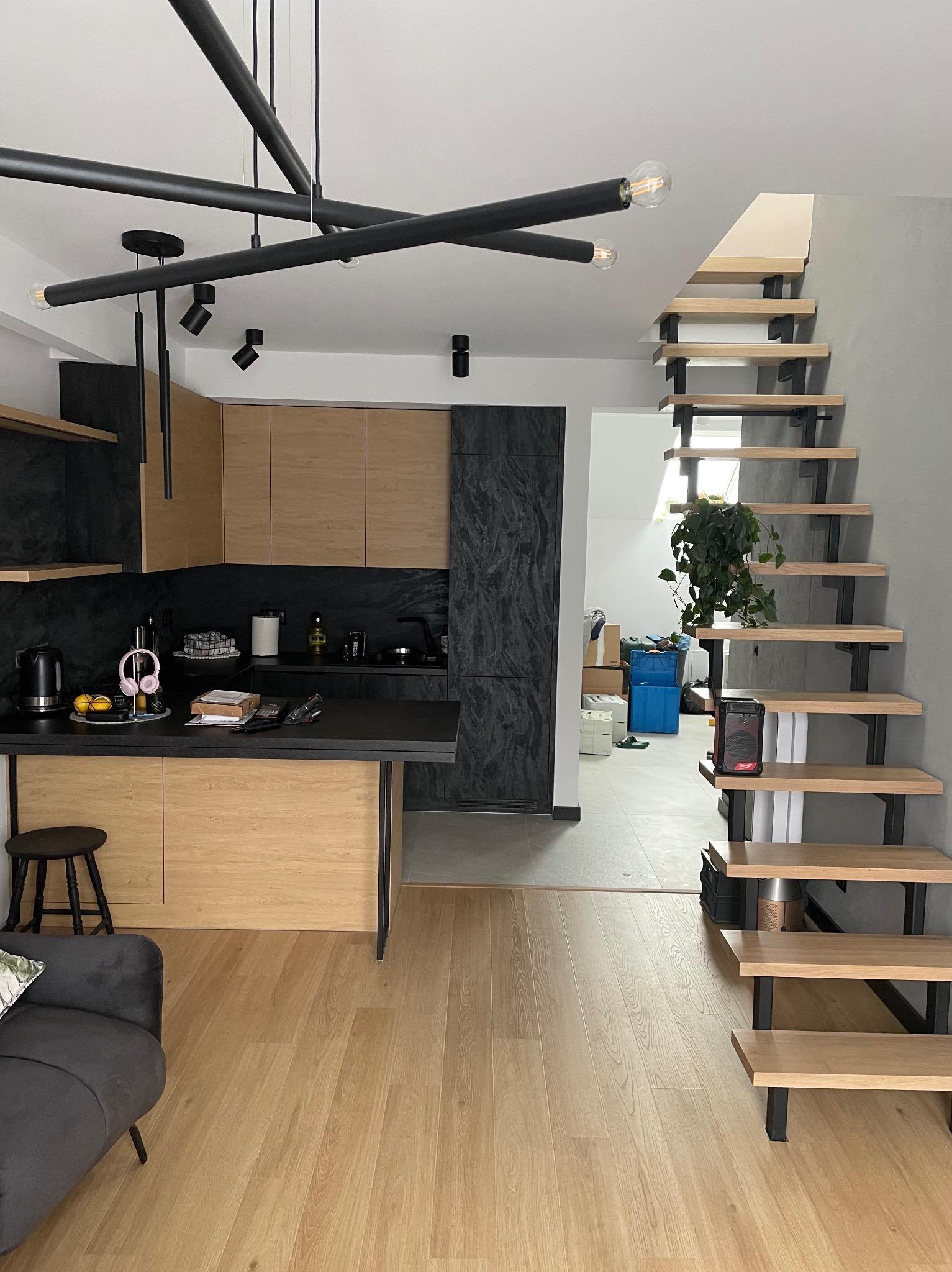 Nowoczesna kuchnia z drewnianymi frontami i czarnymi elementami wykończeniowymi, widok na drewniane schody z metalową konstrukcją, na blacie kuchennym słuchawki i akcesoria.