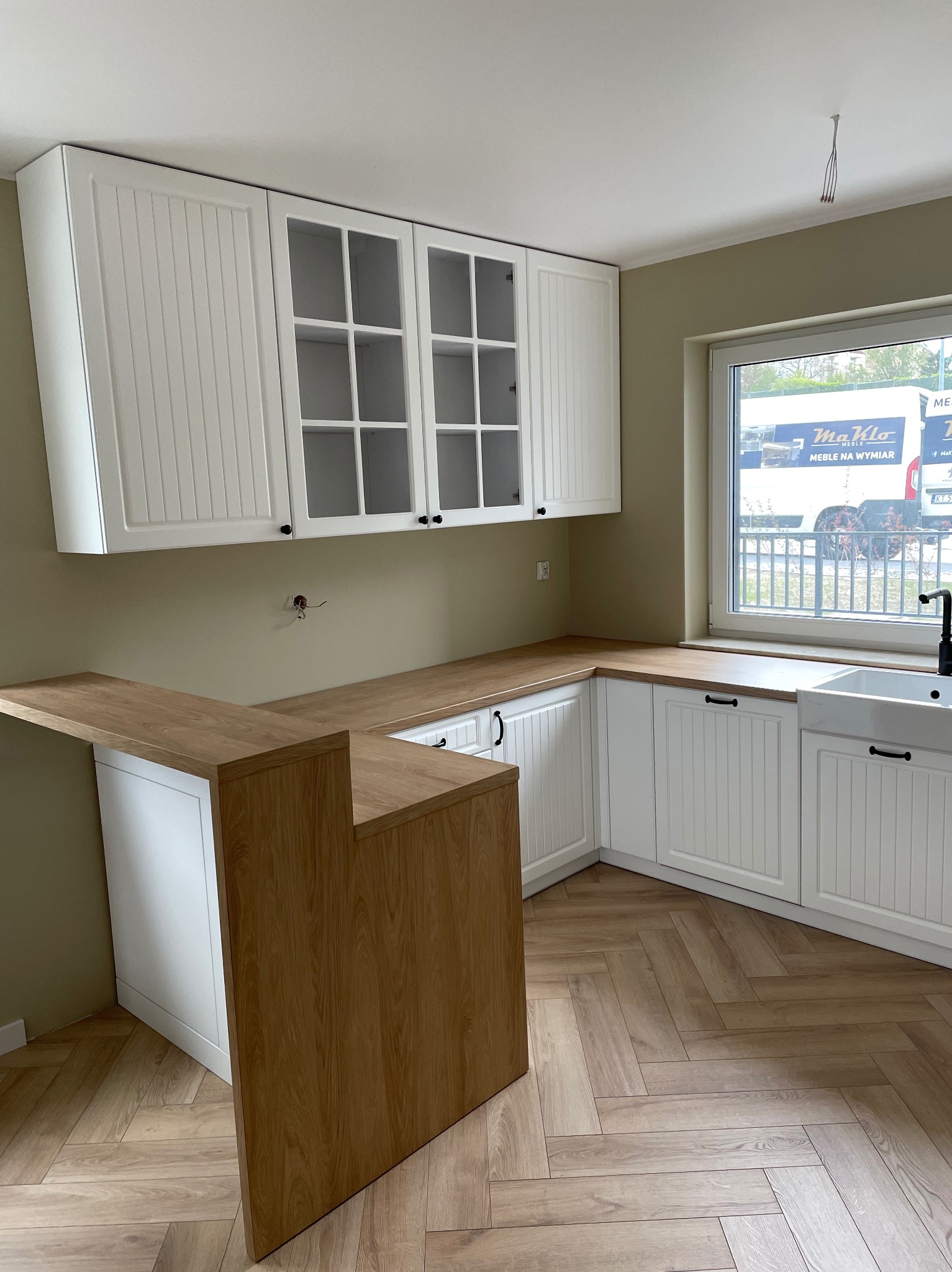 Jasna kuchnia z białymi szafkami z frezowanymi frontami, drewnianym blatem i barkiem, widok na okno z reklamą firmy meblarskiej na samochodzie.