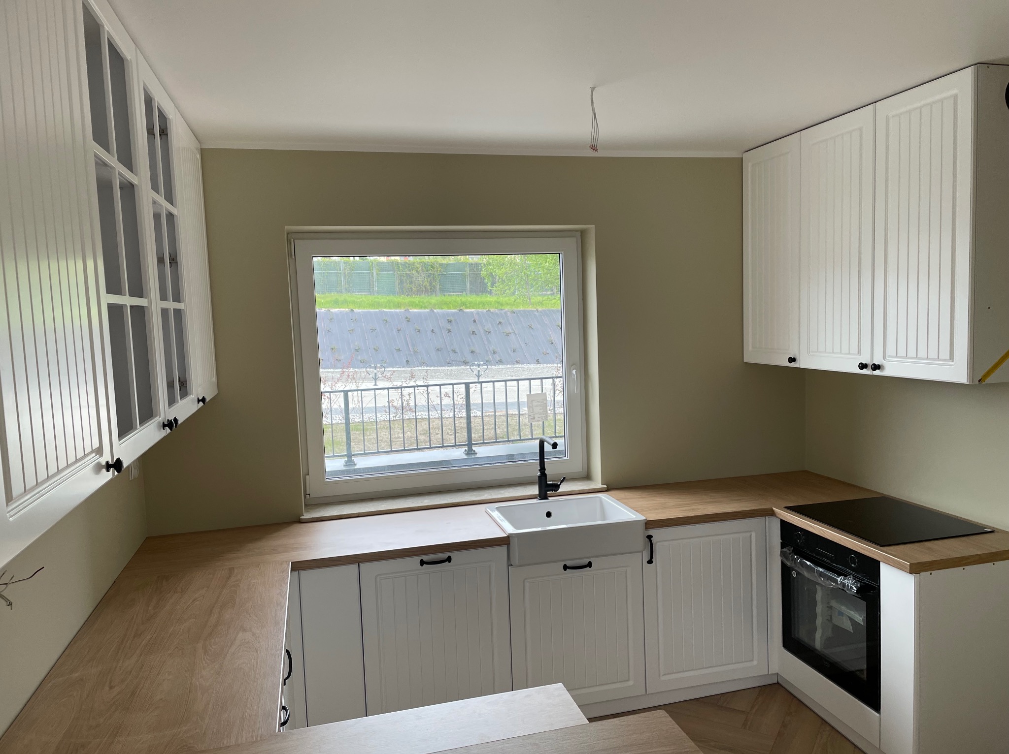 Jasna kuchnia z białymi szafkami w stylu rustykalnym, drewnianym blatem i ceramicznym zlewem farmerskim pod oknem. Widoczne niedokończone podłączenie elektryczne w suficie.