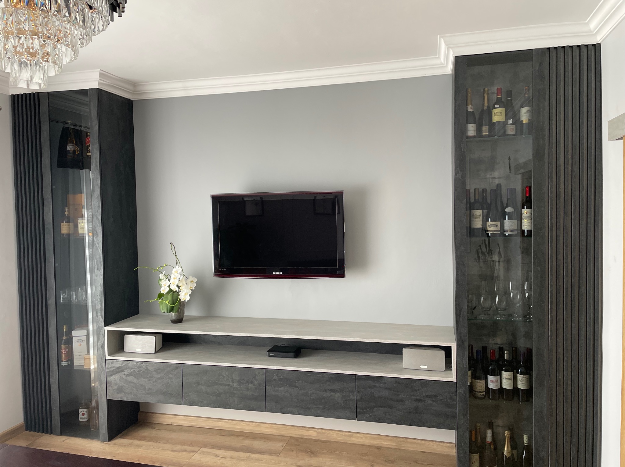 Nowoczesna szafka RTV z szarymi frontami i kamiennym blatem, flankowana przeszklonymi witrynami z kolekcją alkoholi, oświetlona kryształowym żyrandolem.