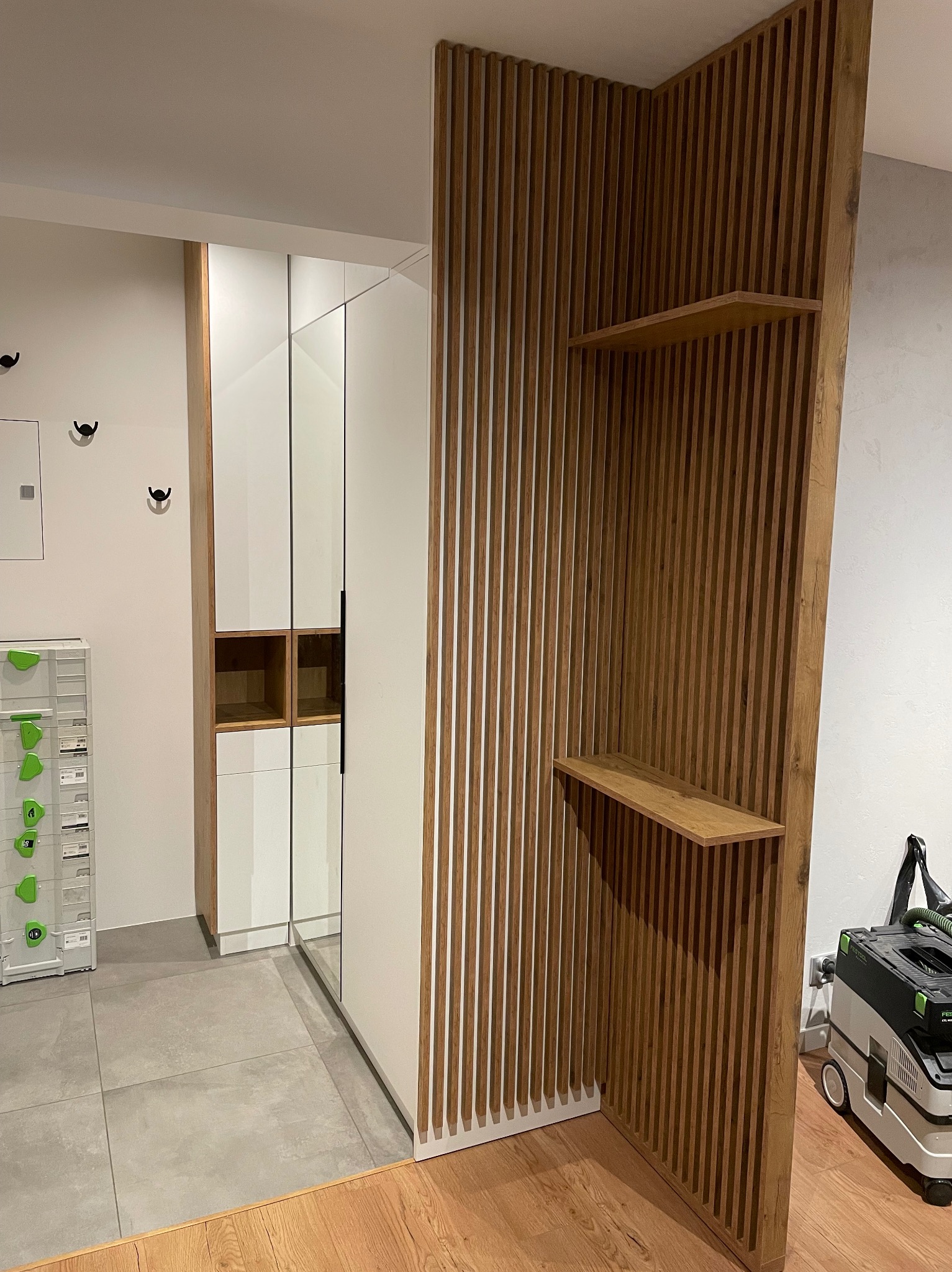 Wnętrze z szafą wnękową z białymi frontami i drewnianą ścianką działową z półkami, widoczny odkurzacz przemysłowy.