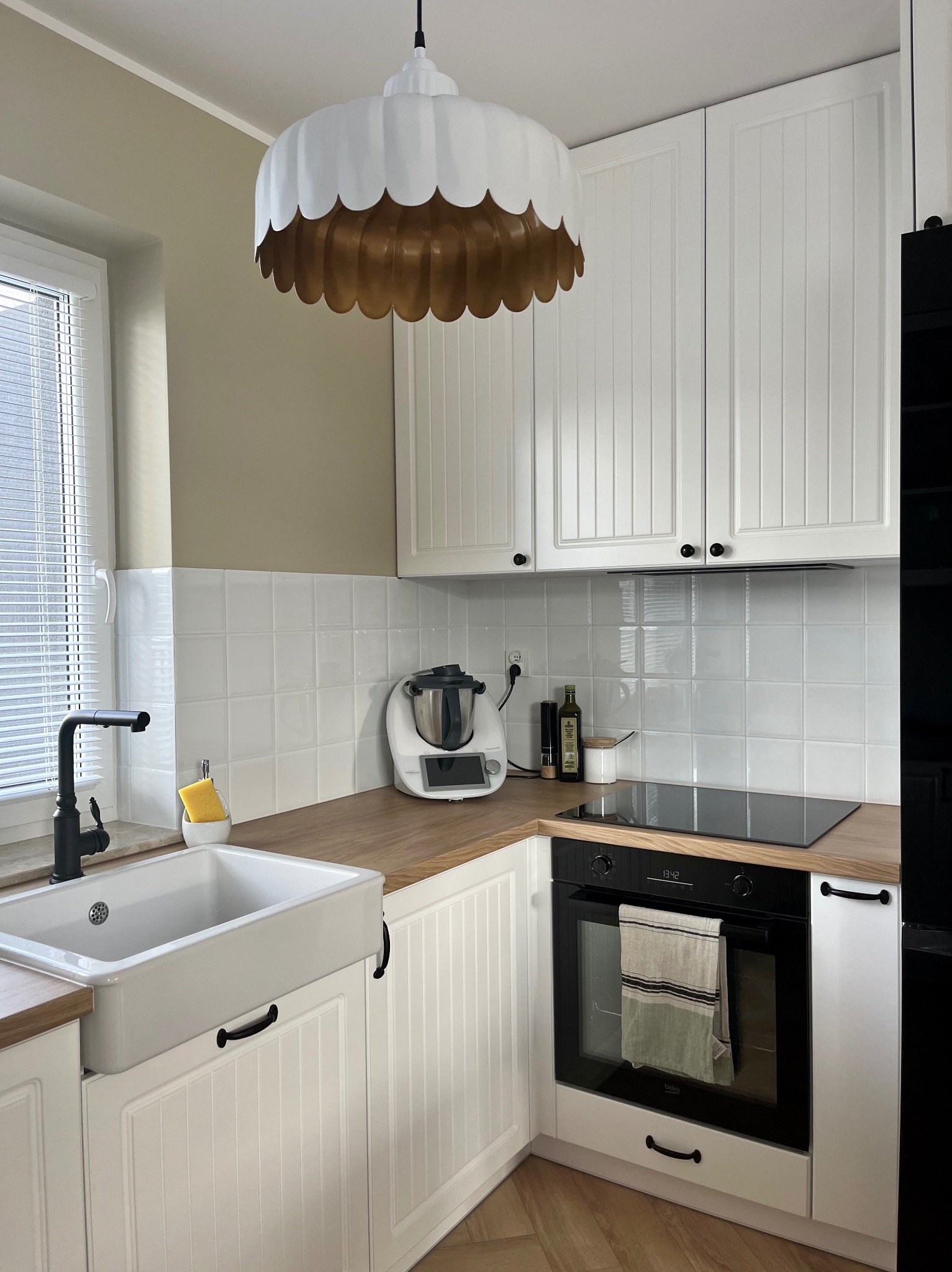 Jasna kuchnia w stylu rustykalnym z białymi szafkami z frezowanymi frontami, drewnianym blatem, ceramicznym zlewem farmerskim i ozdobną lampą w kształcie tortu.