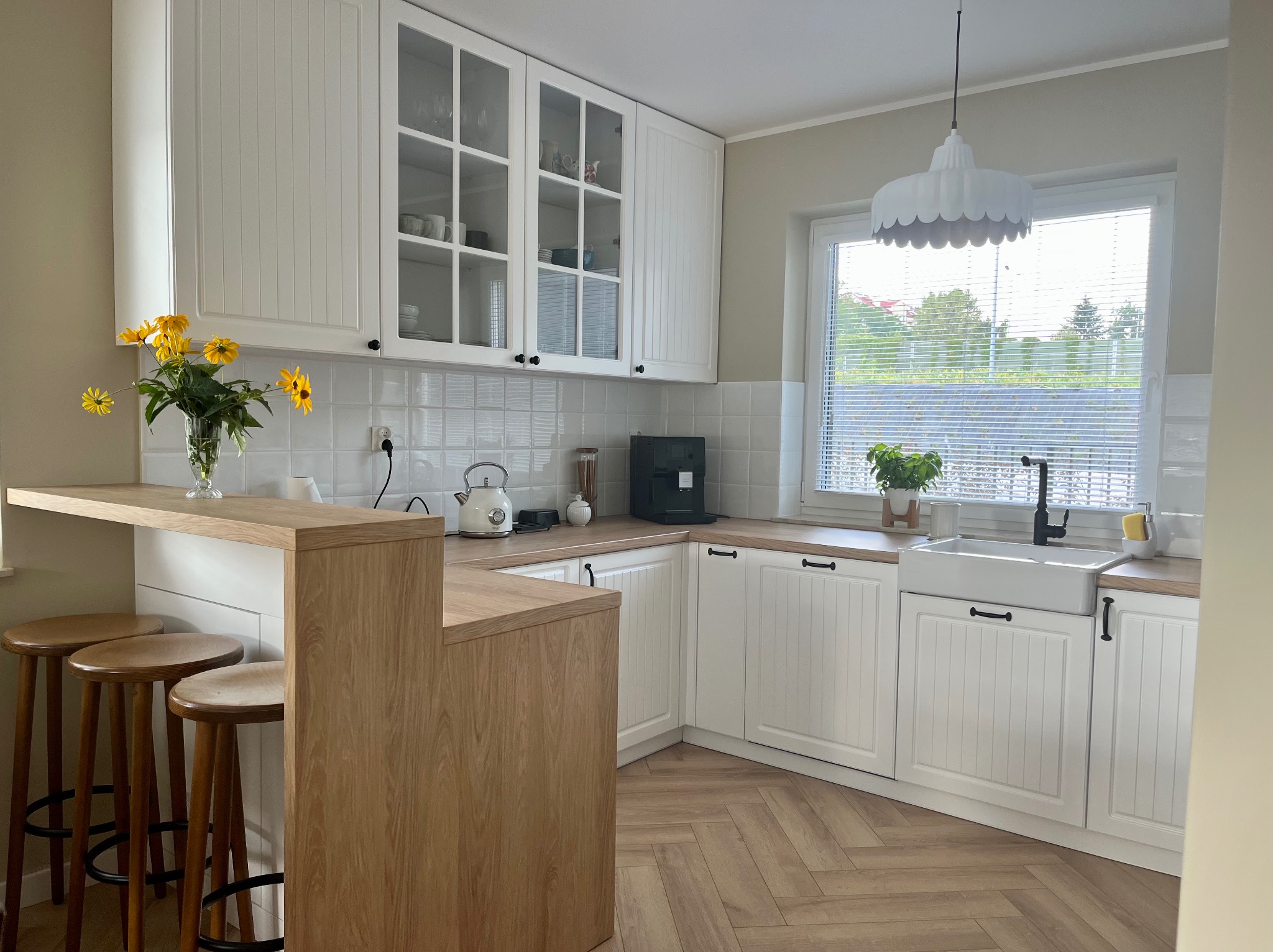 Jasna kuchnia z białymi frontami szafek, drewnianym blatem i barkiem, żółte kwiaty w wazonie, podłoga ułożona w jodełkę, okno z żaluzjami.