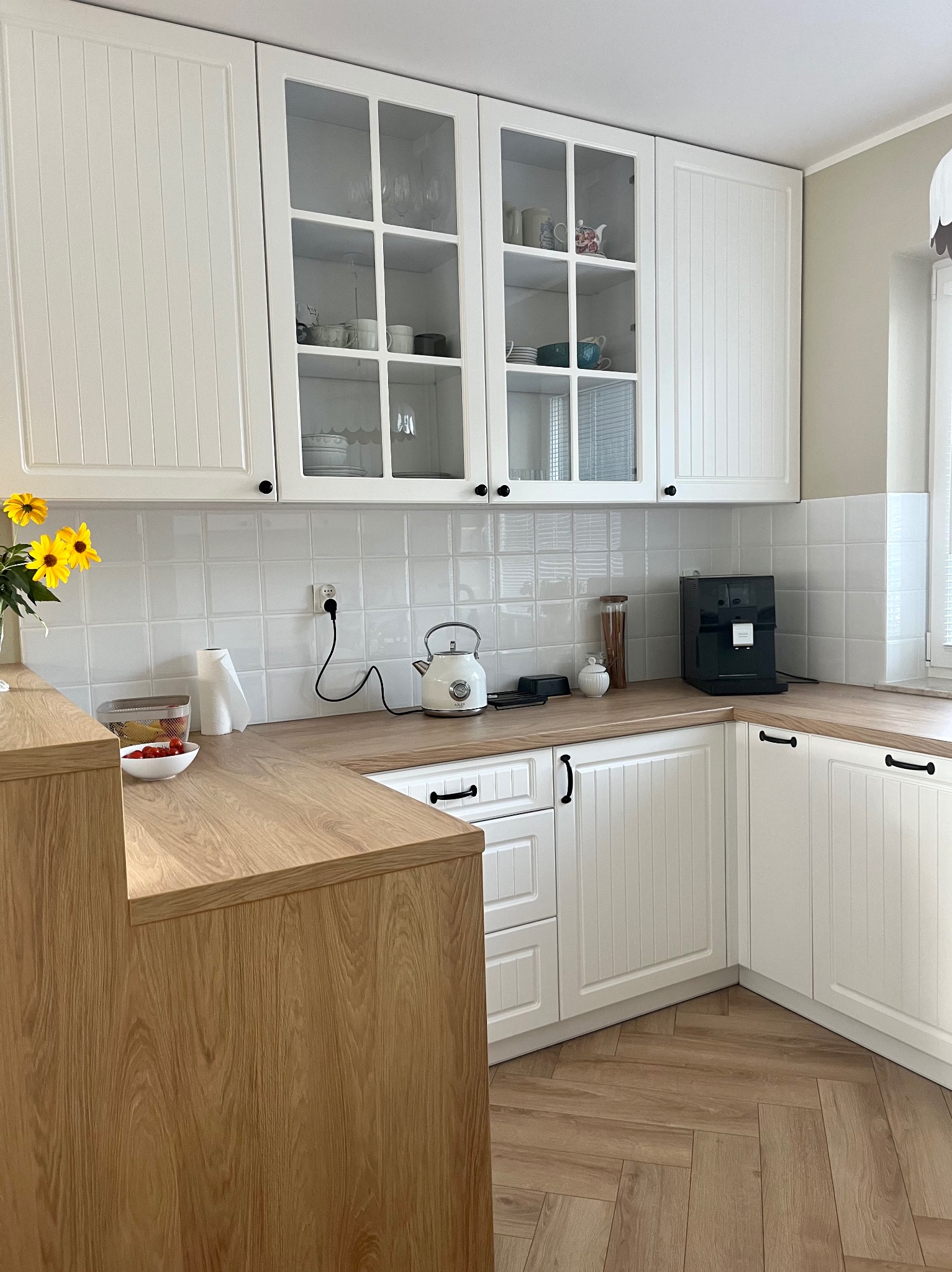 Jasna kuchnia z białymi szafkami z przeszklonymi górnymi frontami, drewnianym blatem i podłogą ułożoną w jodełkę, wyposażona w czarny ekspres do kawy i czajnik.