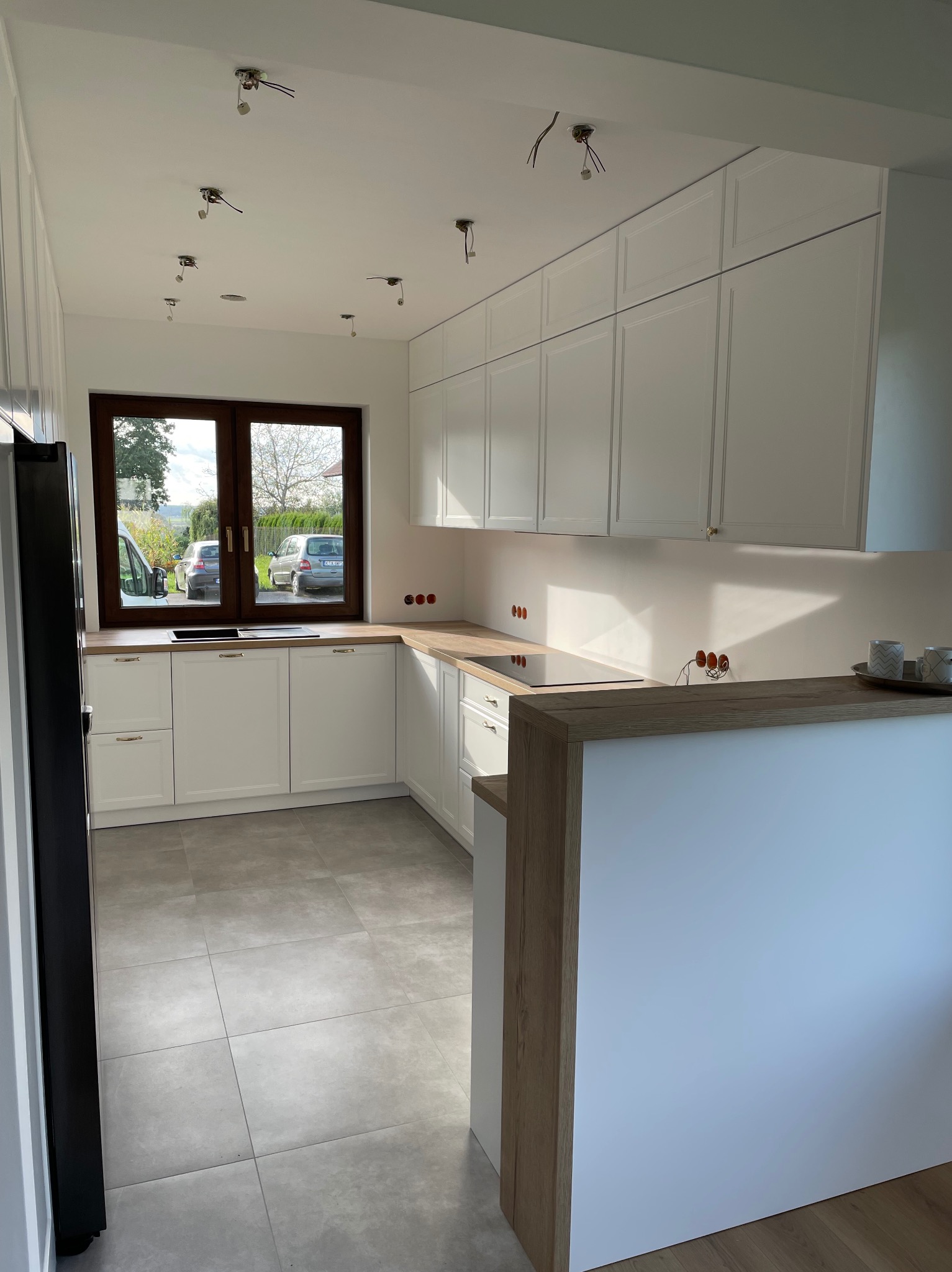 Jasna kuchnia z białymi szafkami i drewnianym blatem, widoczne niedokończone podłączenia elektryczne w suficie i gniazdkach, okno z widokiem na podwórko.