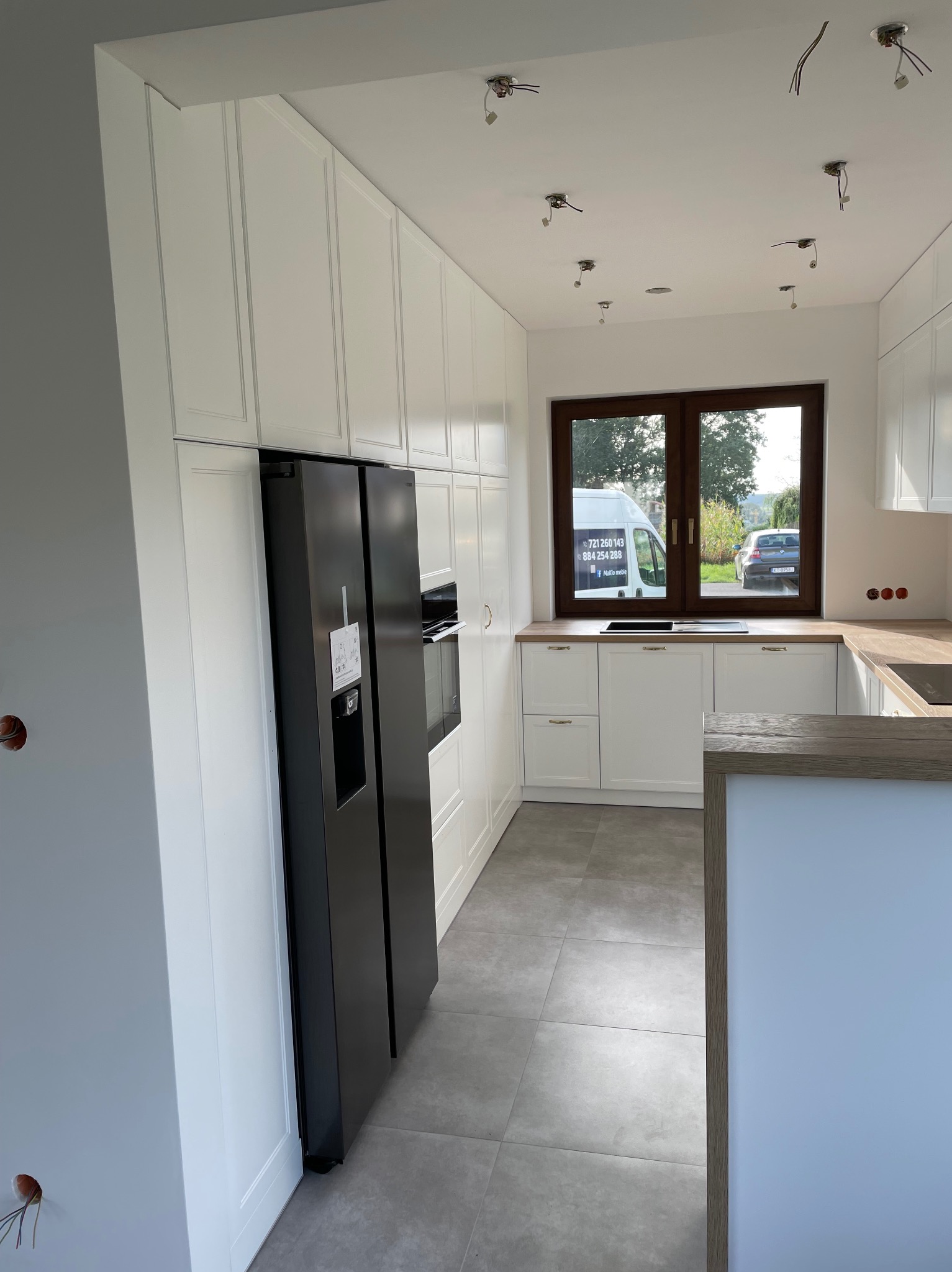 Wnętrze kuchni w trakcie montażu, białe szafki z frezowanymi frontami, czarna lodówka side-by-side, widoczne wystające kable z sufitu, okno z widokiem na samochody.