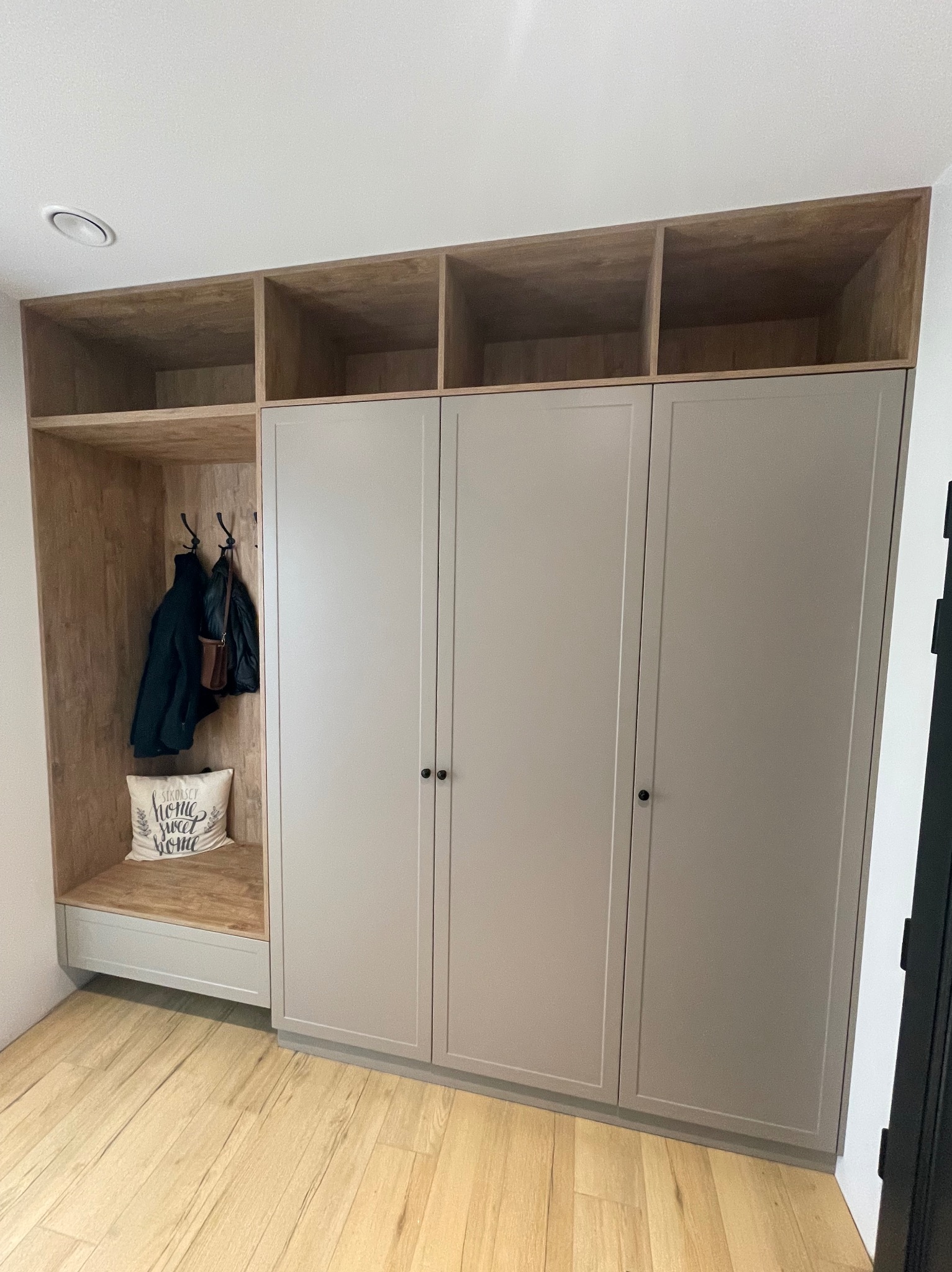 Zabudowa przedpokoju z szafą z szarymi frontami, otwartą wnęką z wieszakami i półką, oraz górnymi półkami z jasnego drewna.
