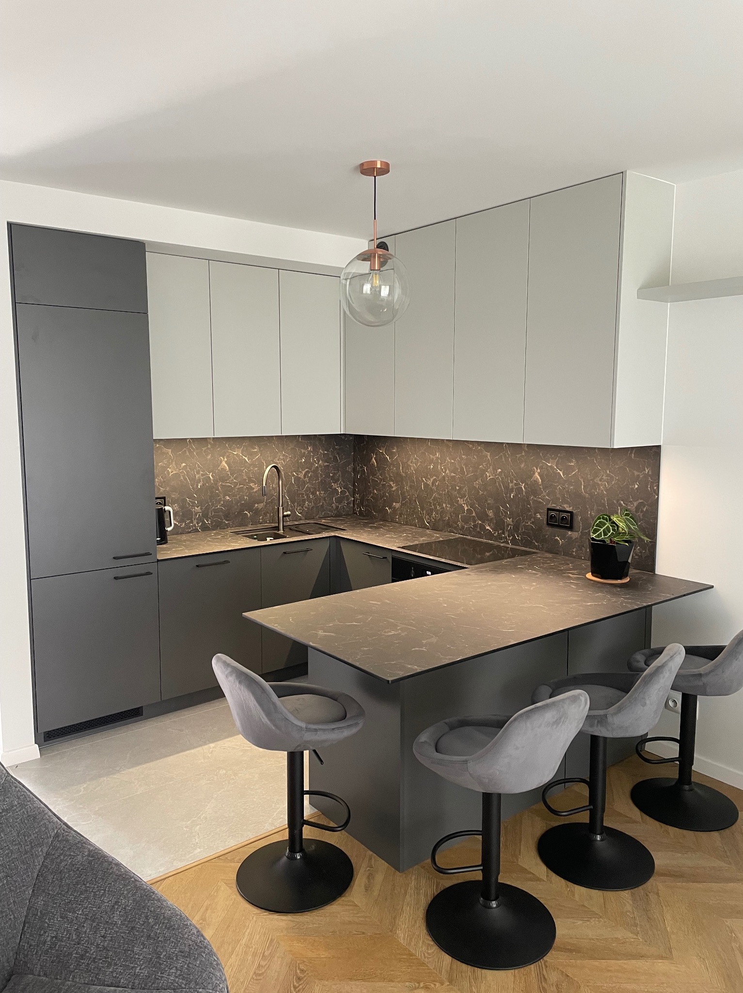 Nowoczesna kuchnia z szarymi i białymi szafkami, blatem imitującym marmur, wyspą z trzema welurowymi hokerami i miedzianą lampą wiszącą.