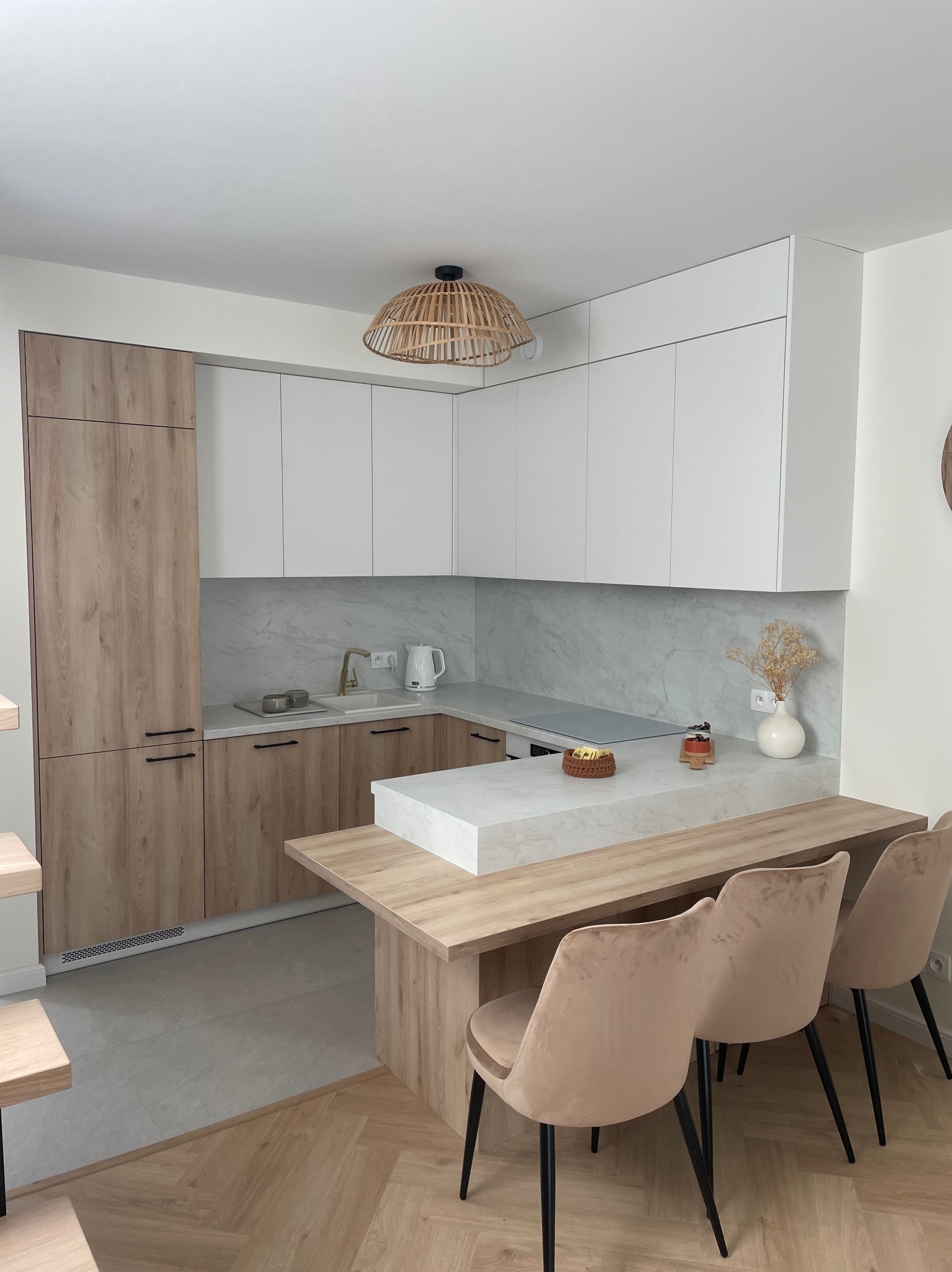 Nowoczesna kuchnia z białymi górnymi szafkami i drewnopodobnymi dolnymi, wyspa z blatem z imitacji marmuru i drewnianym przedłużeniem pełniącym funkcję barku z trzema krzesłami o beżowej tapicerce.