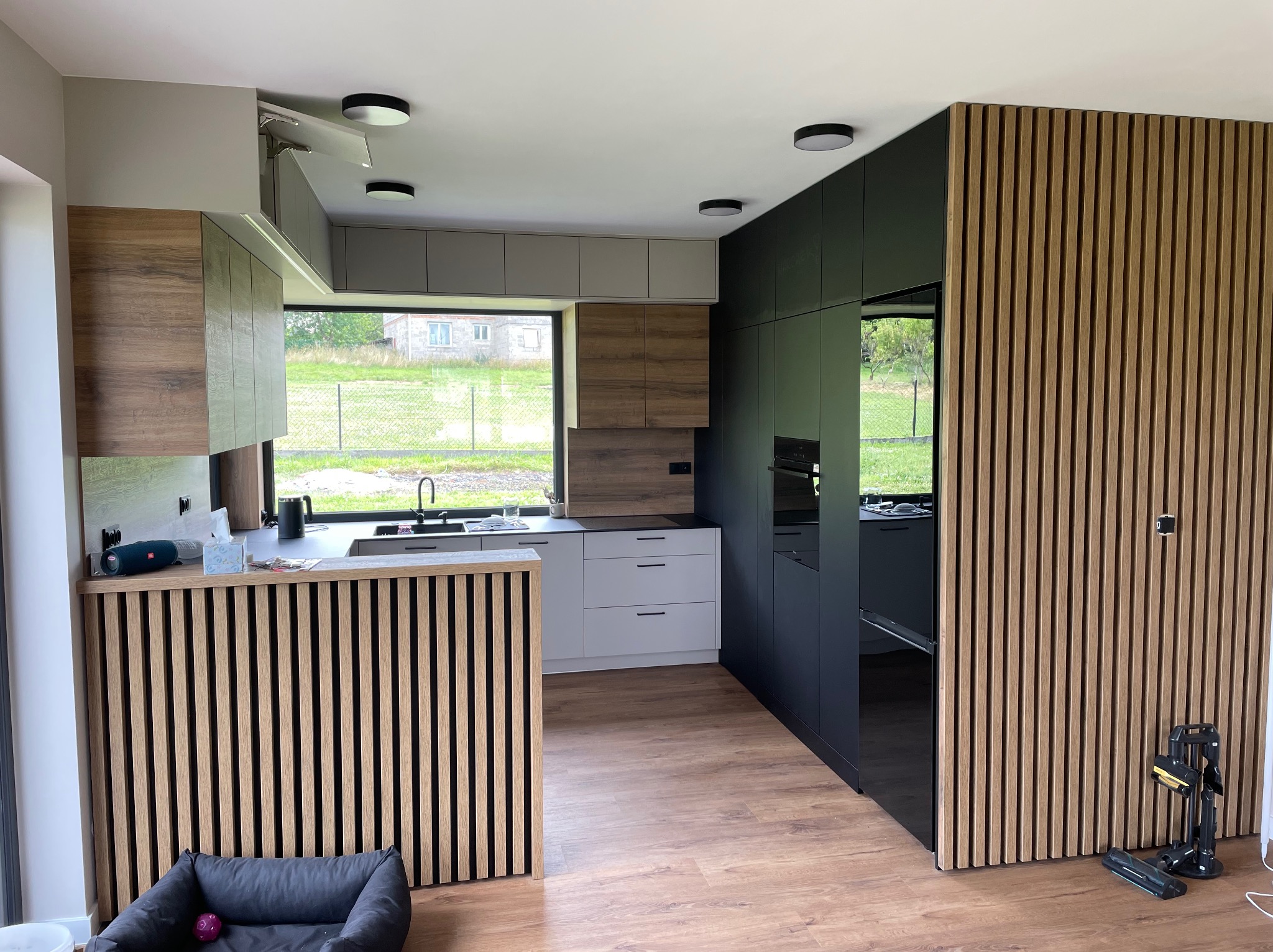 Nowoczesna kuchnia z drewnianymi lamelami, szarymi i drewnopodobnymi szafkami, czarnym sprzętem AGD i oknem z widokiem na ogród. Na podłodze panele imitujące drewno. Widoczne akcesoria kuchenne...