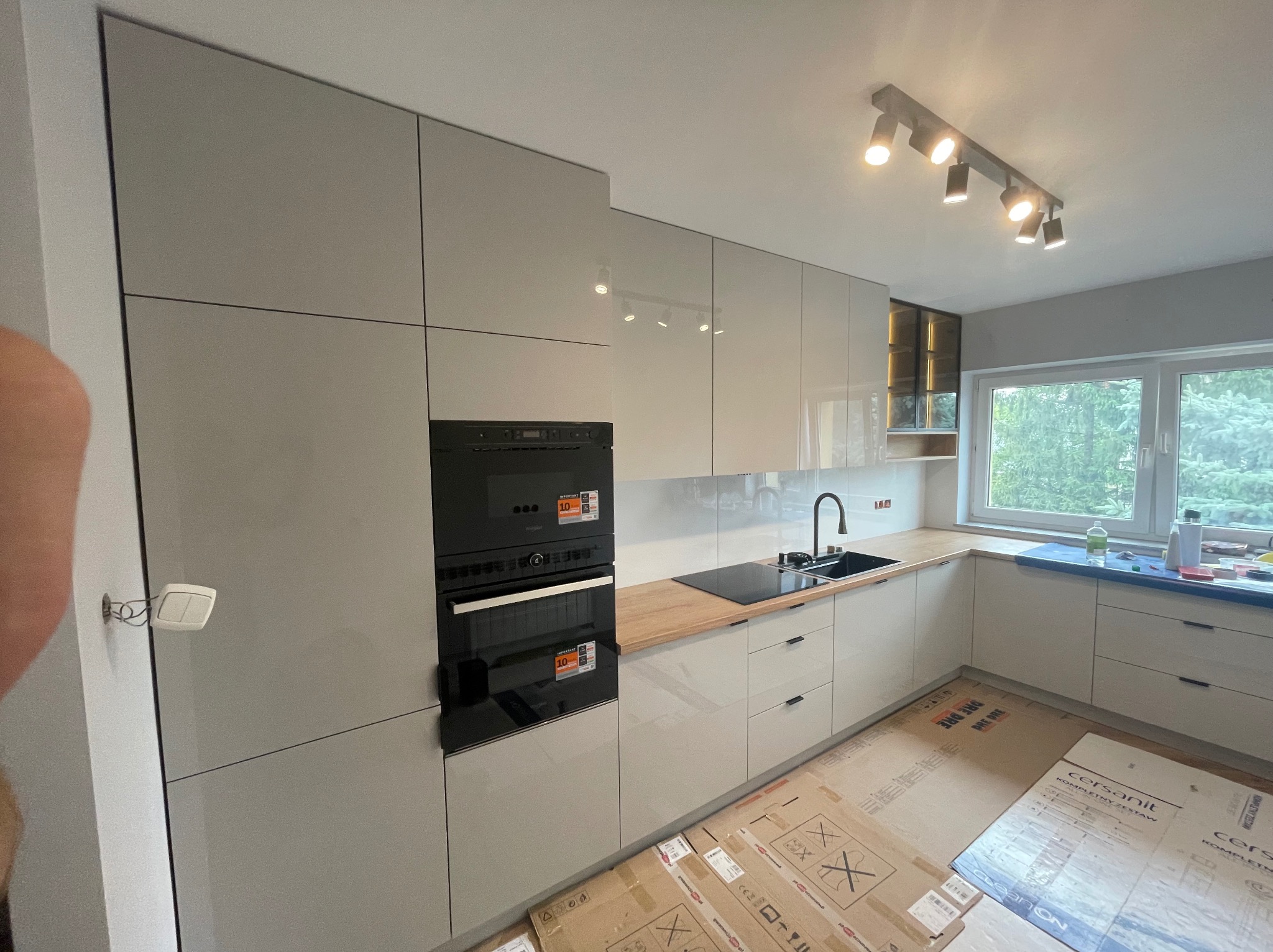 Jasna kuchnia z zabudową na wymiar w trakcie montażu, widoczne kartony na podłodze, minimalistyczny design, czarny piekarnik i płyta grzewcza.