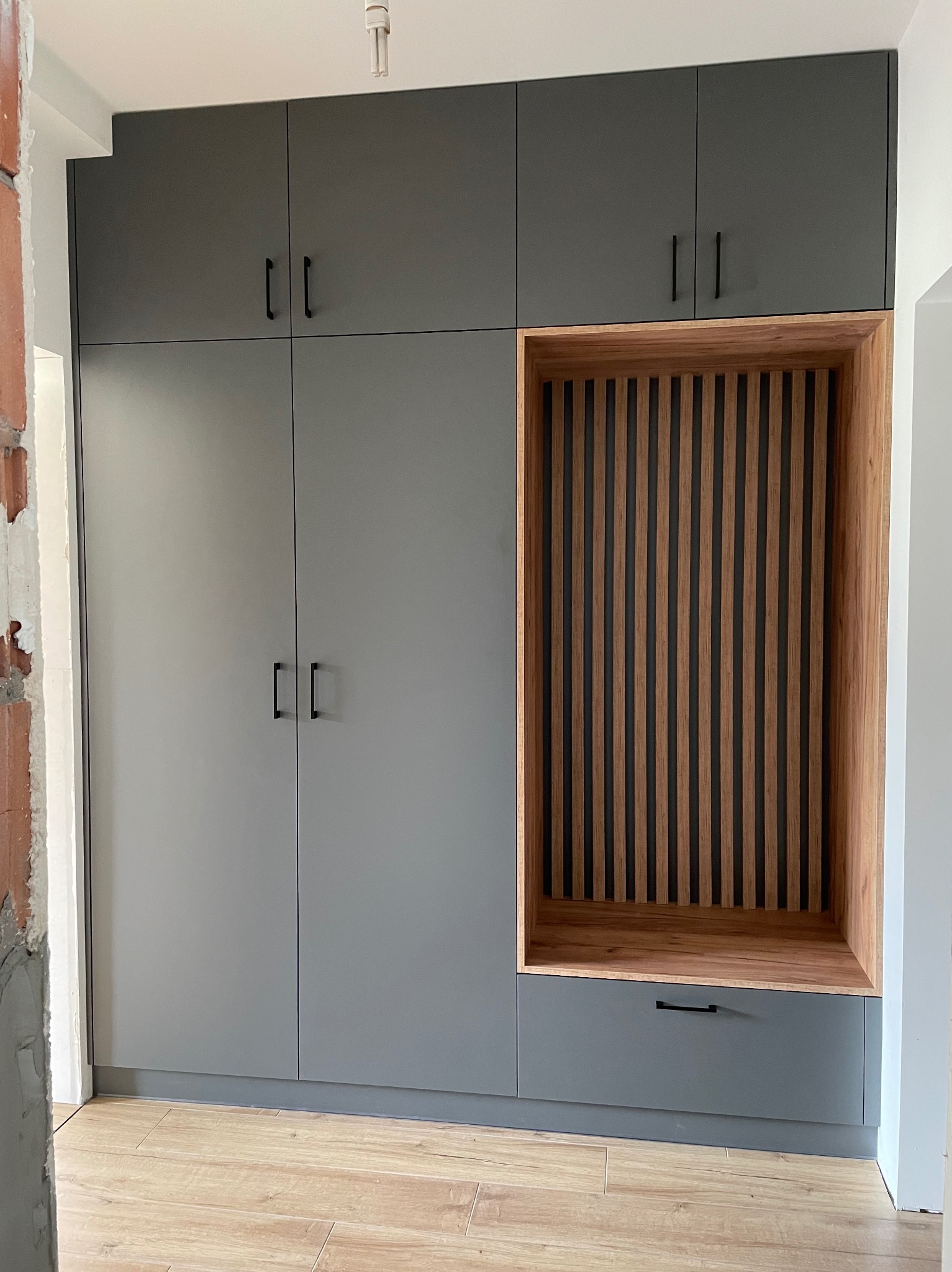 Szara szafa wnękowa z drewnianą wnęką z pionowymi listwami i szufladą, widoczne niedokończone wykończenie ściany.