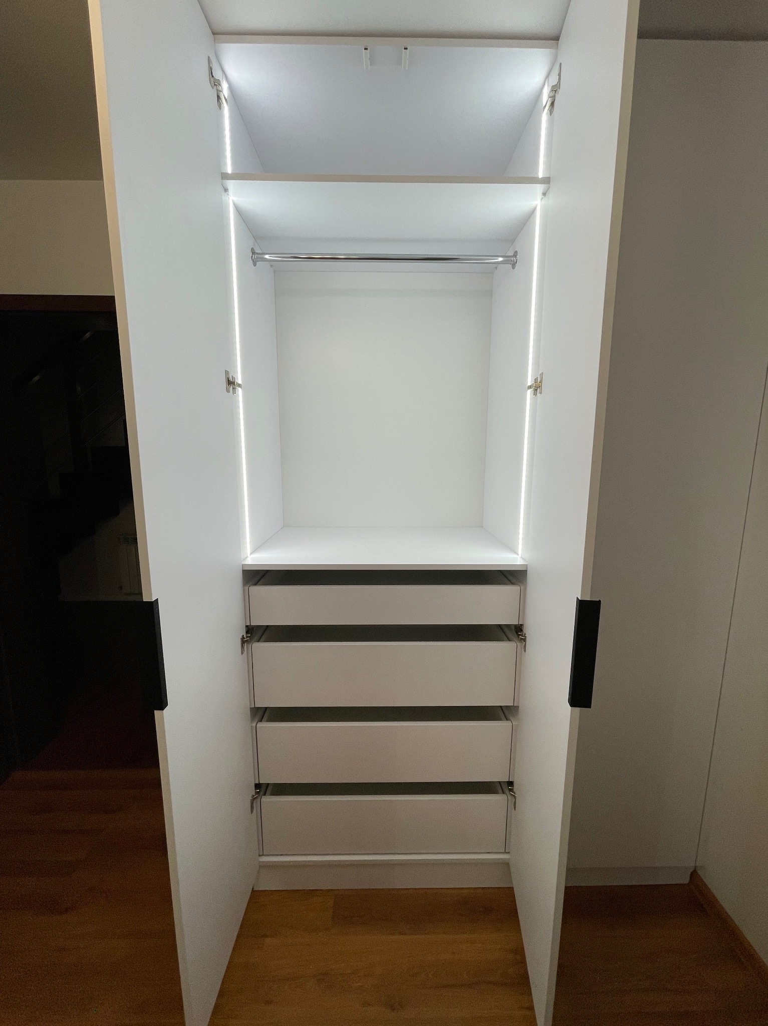 Wnętrze otwartej, białej szafy wnękowej z trzema szufladami, półką i drążkiem na ubrania, oświetlonej listwami LED, zrealizowanej w Tarnowie.