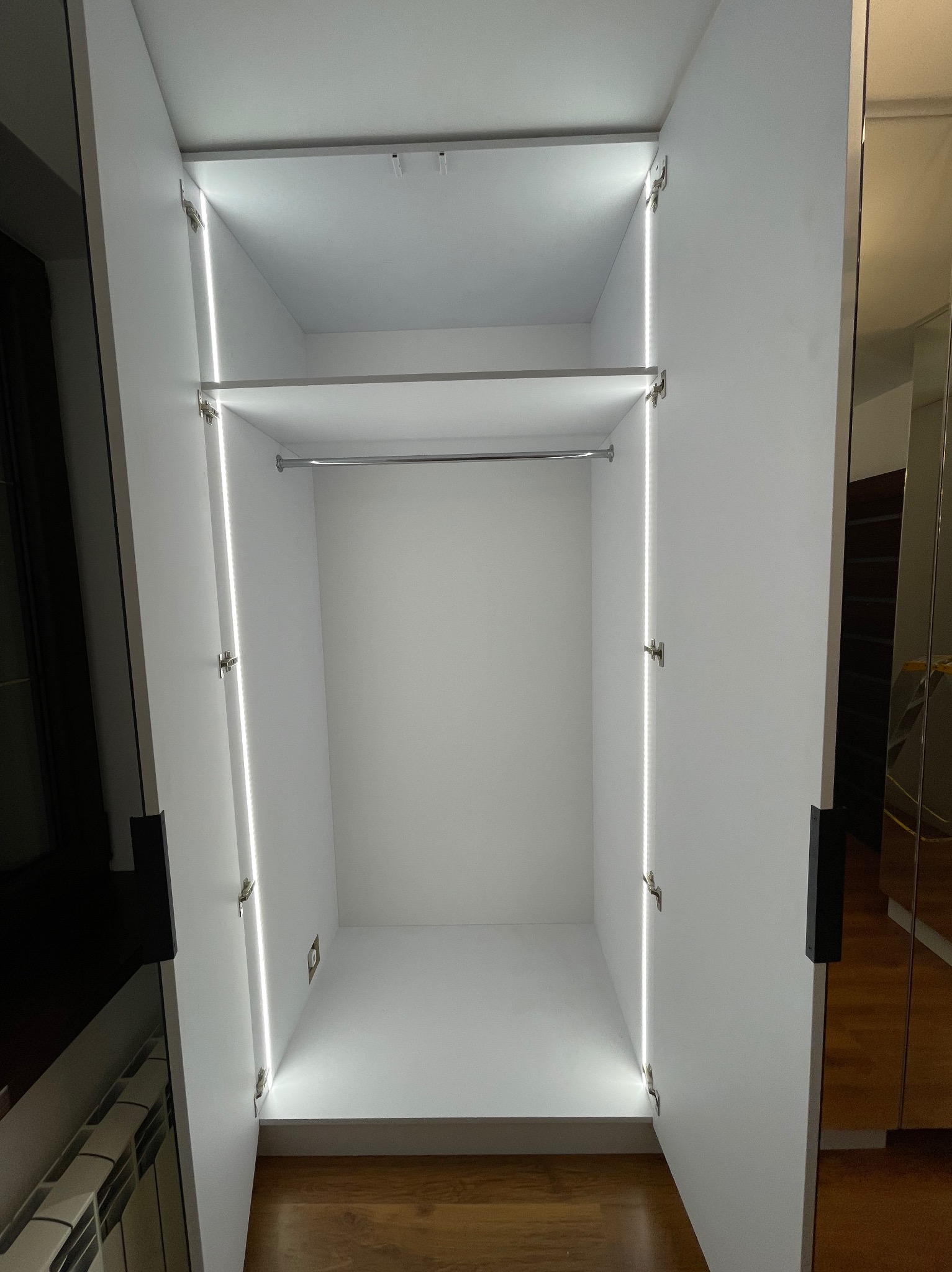 Wnętrze białej szafy wnękowej z otwartymi drzwiami, oświetleniem LED po bokach i drążkiem na ubrania.