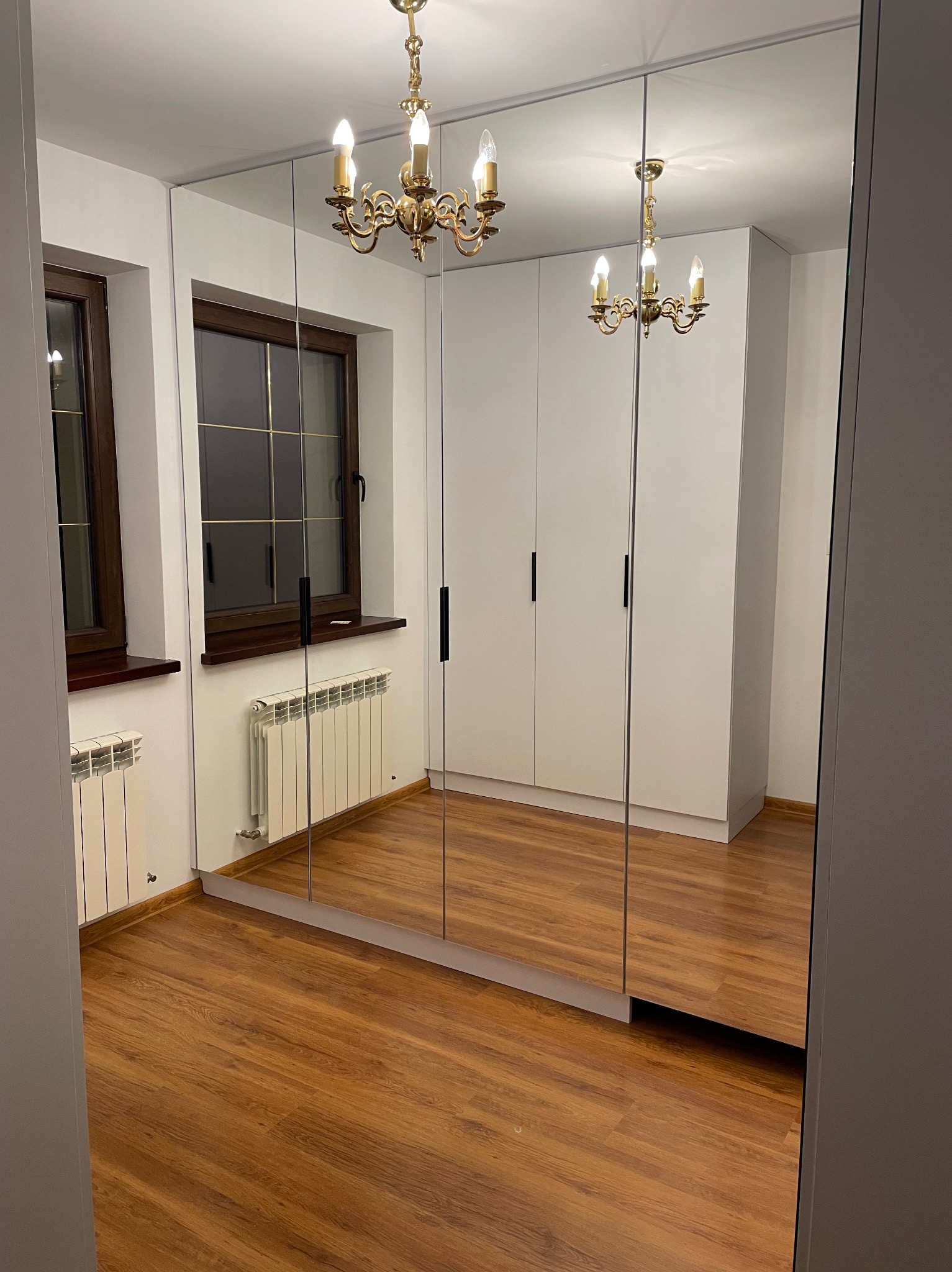 Biała szafa wnękowa z lustrzanymi drzwiami, odbijająca klasyczny żyrandol w pokoju z drewnianą podłogą i oknami z ciemnymi ramami.