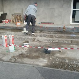 FIRMA BUDOWLANA ROBERT SZCZĄCHOR - Pracownik w trakcie układania ciemnych płytek na schodach wejściowych do budynku z napisem 'ATMOSFERA', widoczne narzędzia i materiały budowlane.