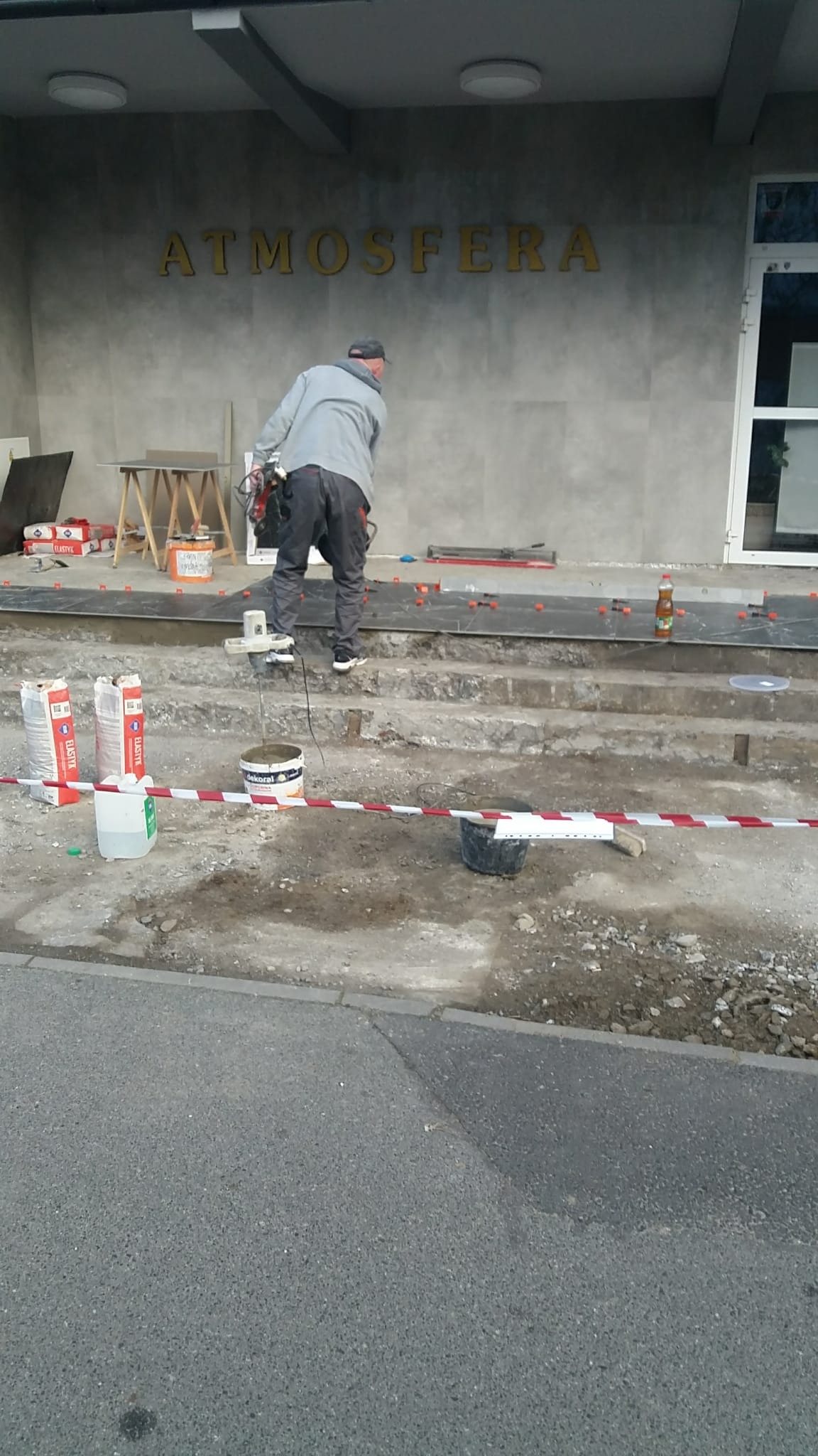 Pracownik w trakcie układania ciemnych płytek na schodach wejściowych do budynku z napisem 'ATMOSFERA', widoczne narzędzia i materiały budowlane.