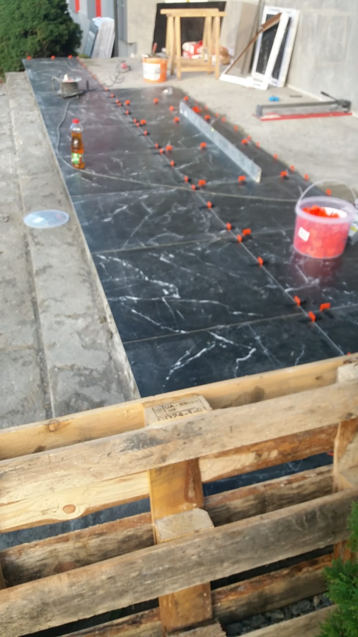 Układanie czarnych płytek imitujących marmur na zewnątrz budynku, widoczne poziomica, narzędzia glazurnicze i butelka z napojem