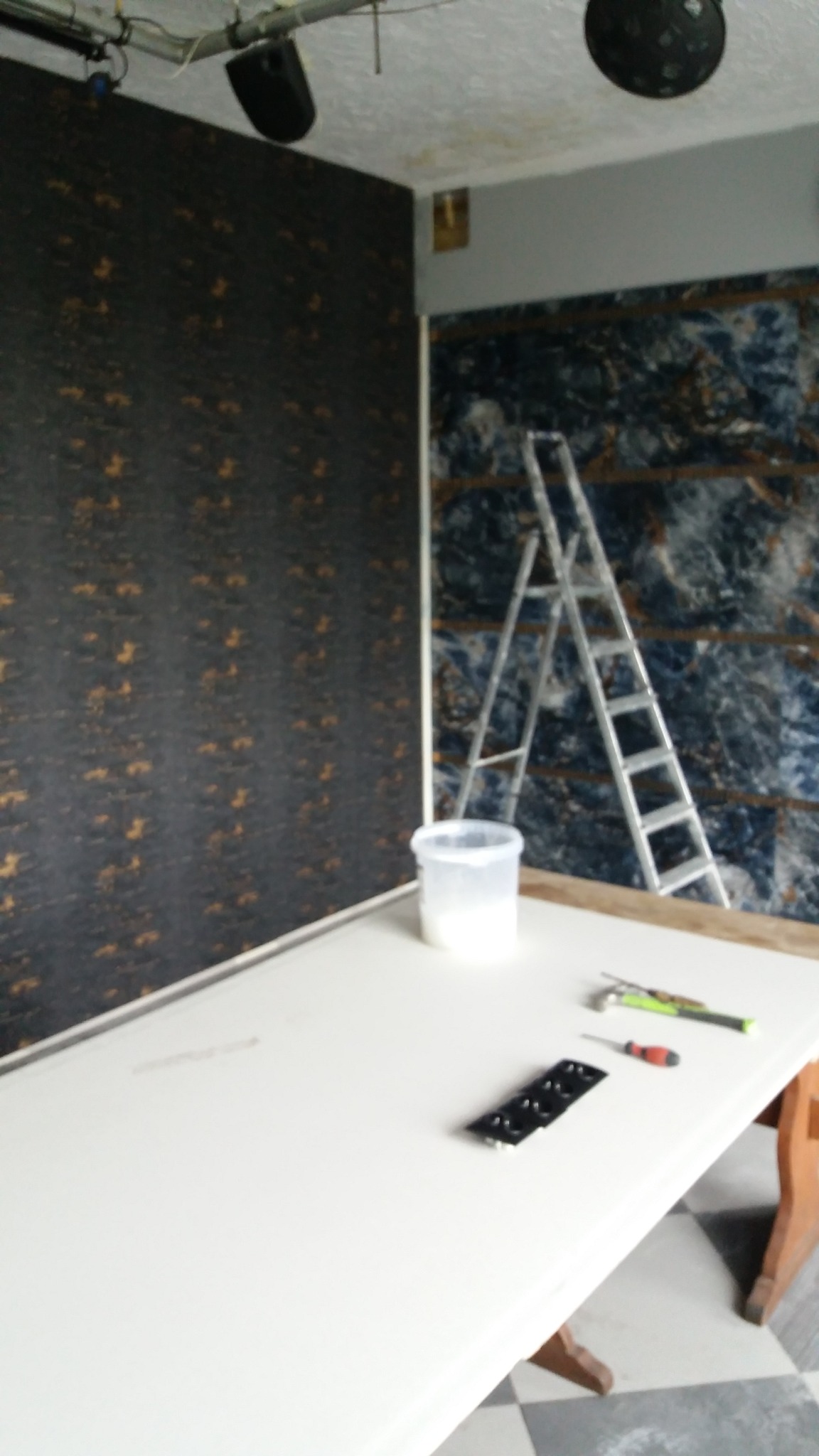 Przygotowania do prac wykończeniowych: ściany z tapetą w odcieniach brązu i błękitu, drabina aluminiowa, wiadro z masą szpachlową, nożyk i włącznik na białym stole.