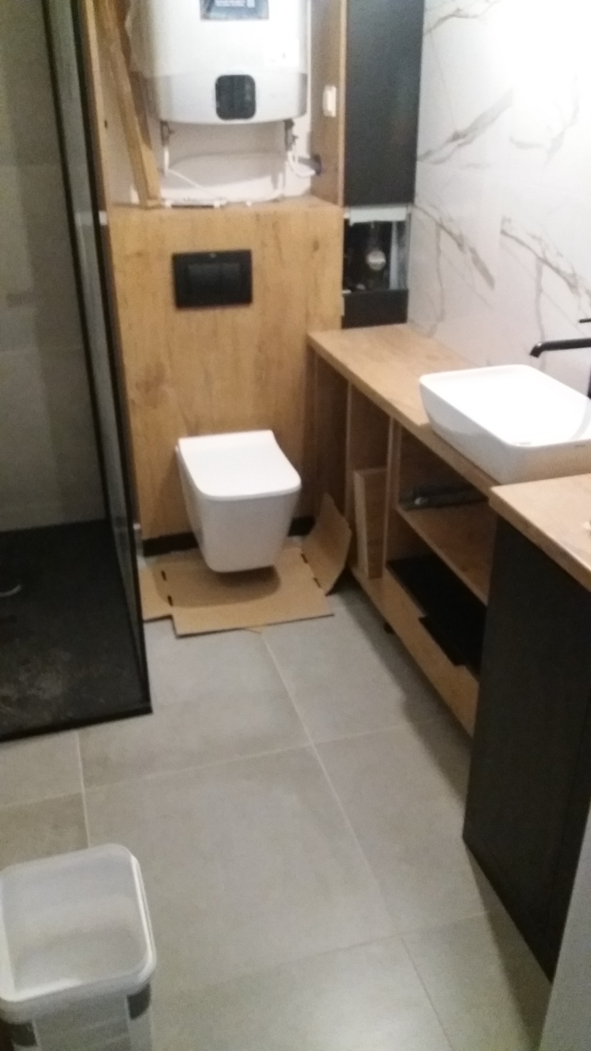 Montaż podwieszanej toalety z zabudową drewnopodobną, widoczny bojler i umywalka na blacie, kartony zabezpieczające podłogę.