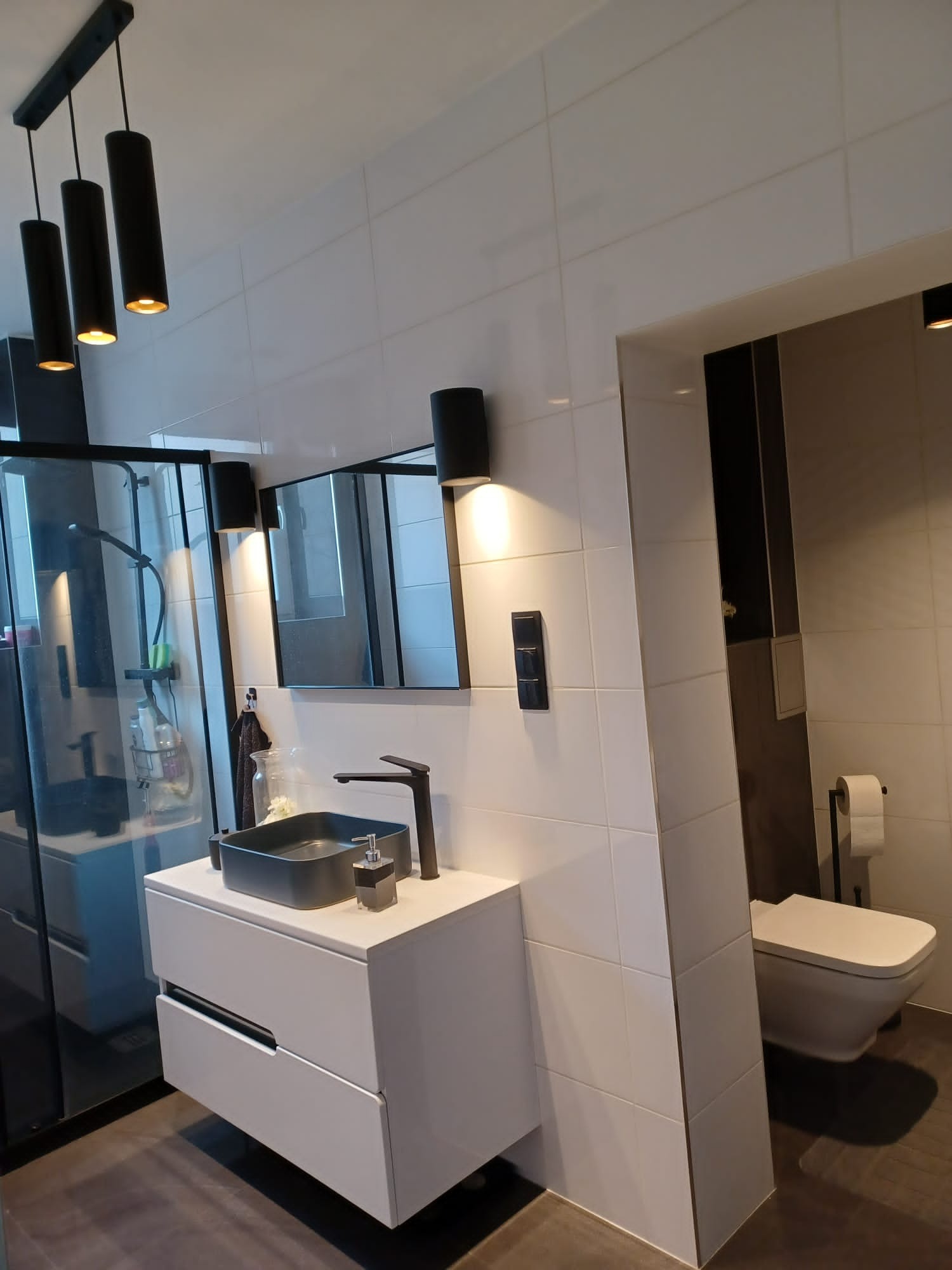 Nowoczesna łazienka z białą armaturą, czarną baterią umywalkową i oświetleniem punktowym, widok na kabinę prysznicową z ciemną ramą i toaletę podwieszaną.