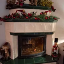 Ozdobny, kaflowy piec kominkowy z paleniskiem w stylu retro, udekorowany świątecznymi figurkami Mikołajów, skrzatów i girlandą z czerwonych bombek i gałązek choinki, z płonącym ogniem wewnątrz.