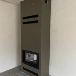 ZAKĄTEK SPÓŁKA Z OGRANICZONĄ ODPOWIEDZIALNOŚCIĄ - Wysoki, wąski kominek w obudowie w kolorze khaki z poziomymi szczelinami wentylacyjnymi umieszczony w rogu surowego pomieszczenia z niepomalowanymi ścianami i betonową podłogą.