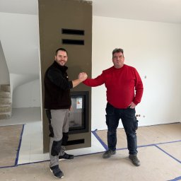ZAKĄTEK SPÓŁKA Z OGRANICZONĄ ODPOWIEDZIALNOŚCIĄ - Dwóch mężczyzn w odzieży roboczej ściska sobie dłonie obok nowoczesnego kominka z wkładem, w surowym wnętrzu z widocznymi schodami w tle.