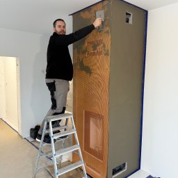 ZAKĄTEK SPÓŁKA Z OGRANICZONĄ ODPOWIEDZIALNOŚCIĄ - Mężczyzna na drabinie, zacierający szarą zaprawą obudowę kominka z kratką wzmacniającą, zabezpieczoną niebieską taśmą malarską.