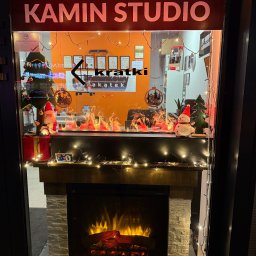 ZAKĄTEK SPÓŁKA Z OGRANICZONĄ ODPOWIEDZIALNOŚCIĄ - Wystawa salonu kominkowego z elektrycznym kominkiem udekorowanym świątecznymi ozdobami, w tle widoczne certyfikaty i logo firmy.