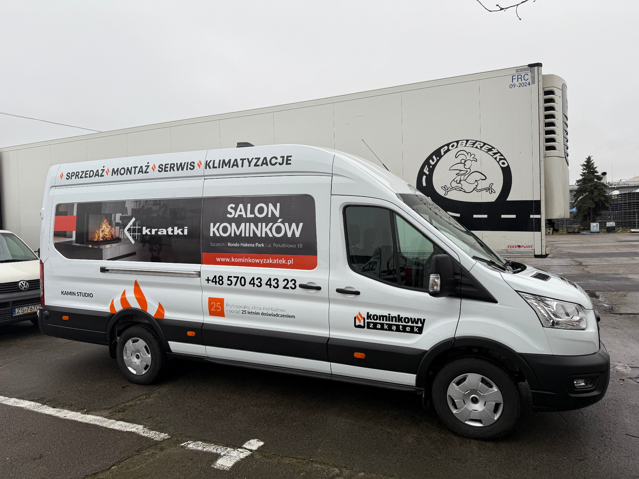 Biały samochód dostawczy z reklamą firmy 'Salon Kominków' z grafiką płonącego kominka w oknie i logiem firmy 'Kominkowy Zakątek'.