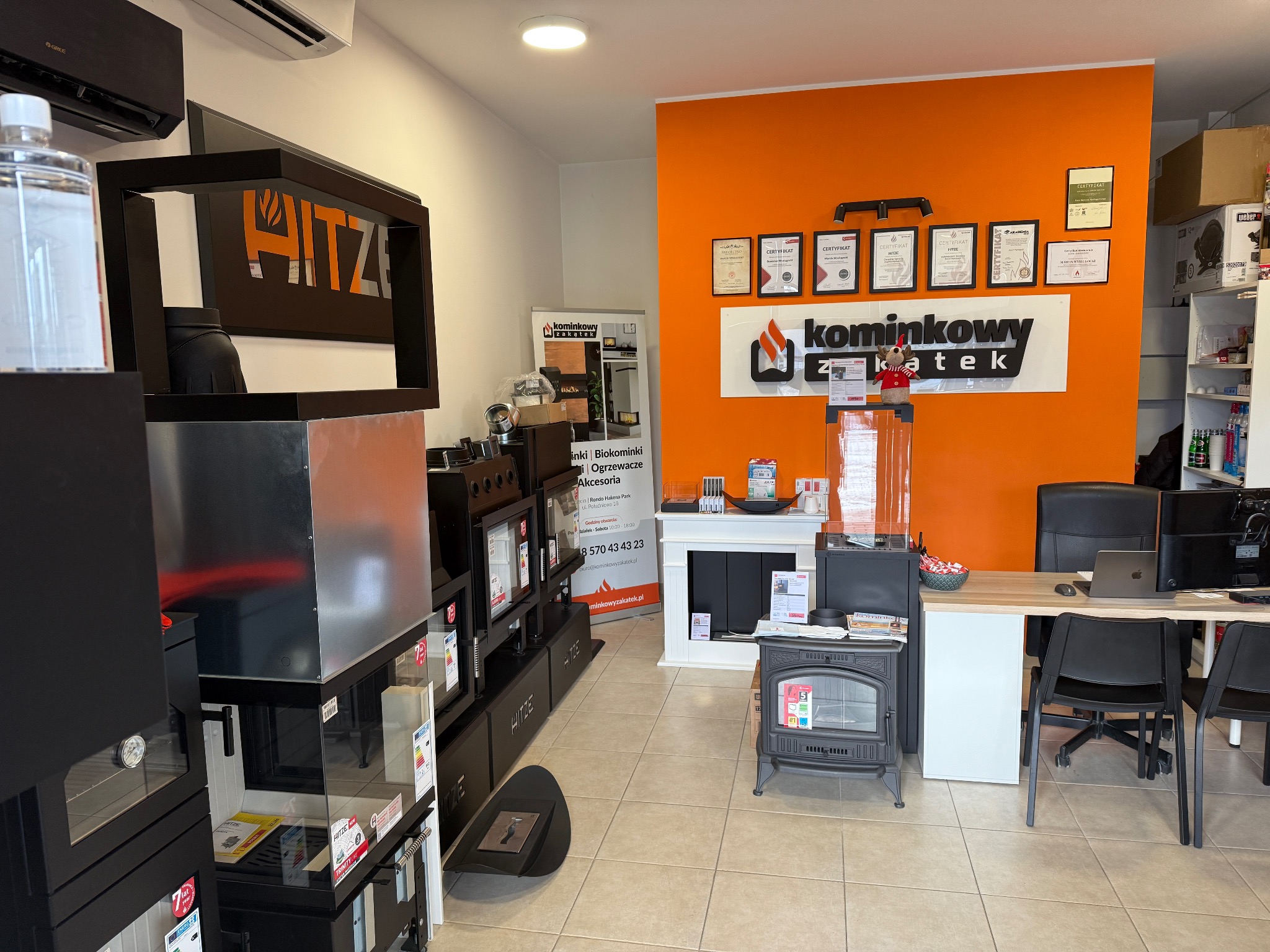 Ekspozycja pieców kominkowych i wkładów kominkowych w salonie sprzedaży, widoczne certyfikaty na pomarańczowej ścianie i logo producenta Hitz.