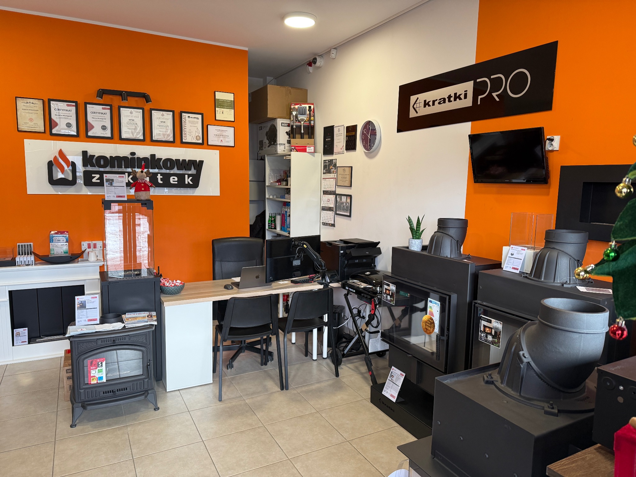 Wnętrze salonu z ekspozycją pieców kominkowych i wkładów kominkowych, na ścianie certyfikaty i logo firmy 'kominkowy z ktek' oraz 'kratki PRO', biurko z laptopem.