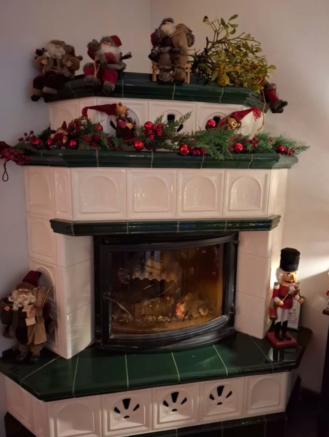 Ozdobny, kaflowy piec kominkowy z paleniskiem w stylu retro, udekorowany świątecznymi figurkami Mikołajów, skrzatów i girlandą z czerwonych bombek i gałązek choinki, z płonącym ogniem wewnątrz.