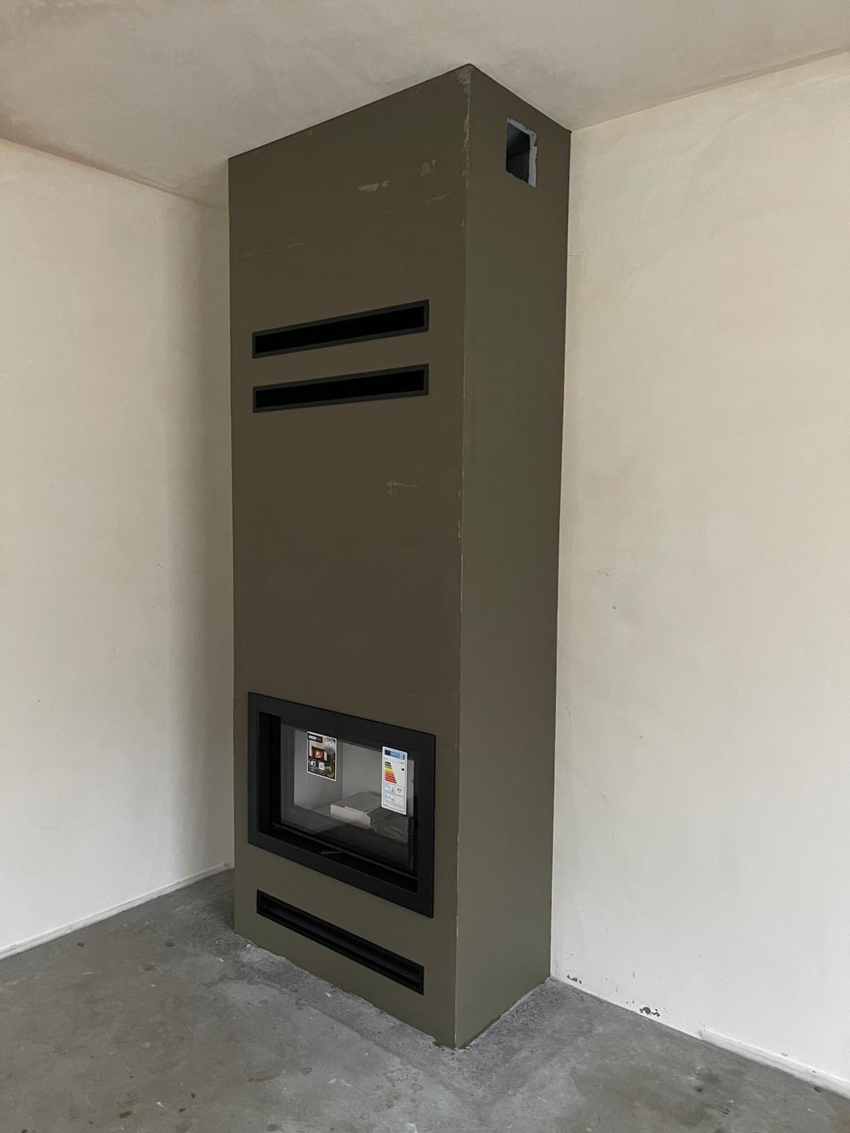 Wysoki, wąski kominek w obudowie w kolorze khaki z poziomymi szczelinami wentylacyjnymi umieszczony w rogu surowego pomieszczenia z niepomalowanymi ścianami i betonową podłogą.