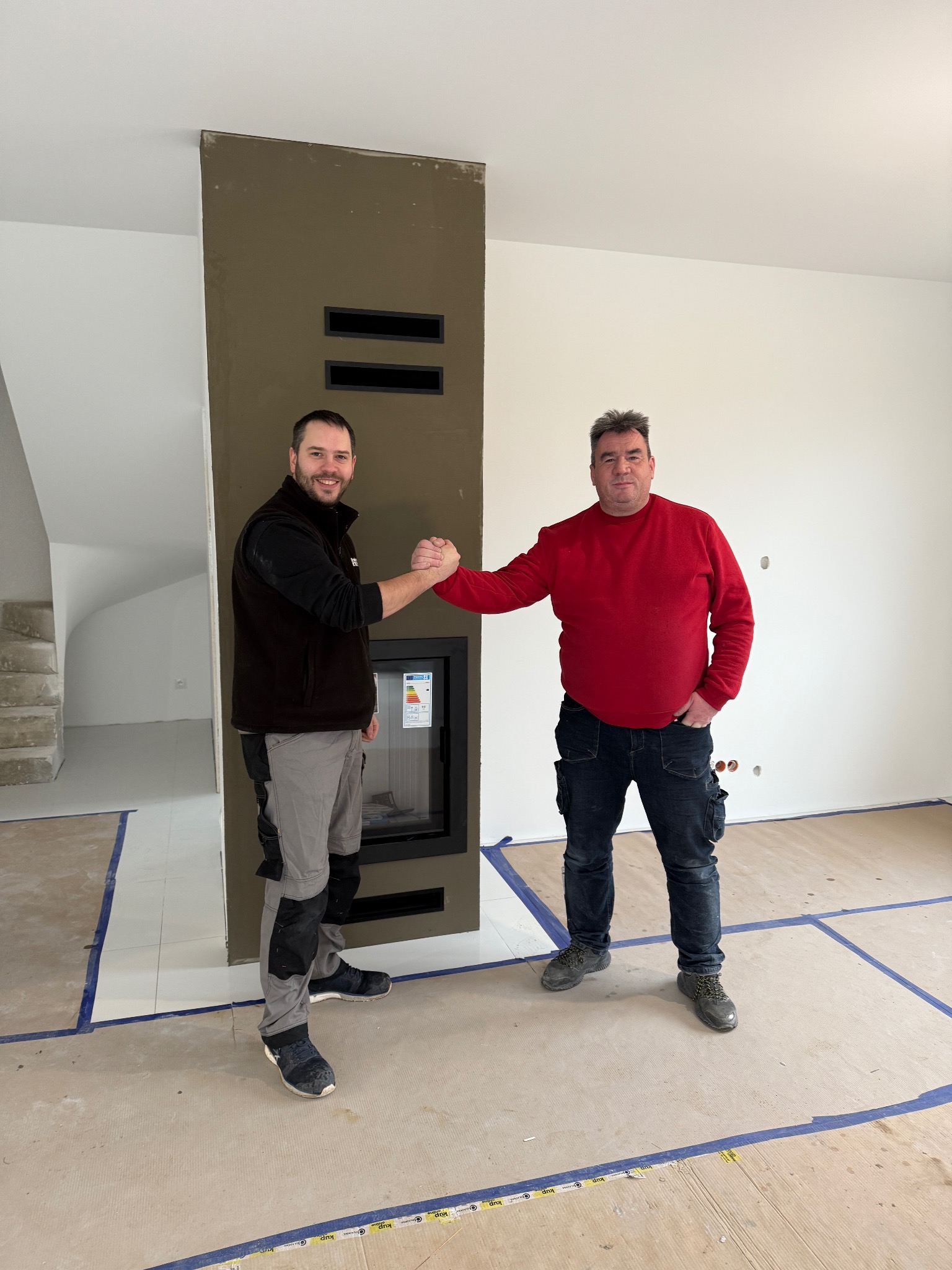 Dwóch mężczyzn w odzieży roboczej ściska sobie dłonie obok nowoczesnego kominka z wkładem, w surowym wnętrzu z widocznymi schodami w tle.