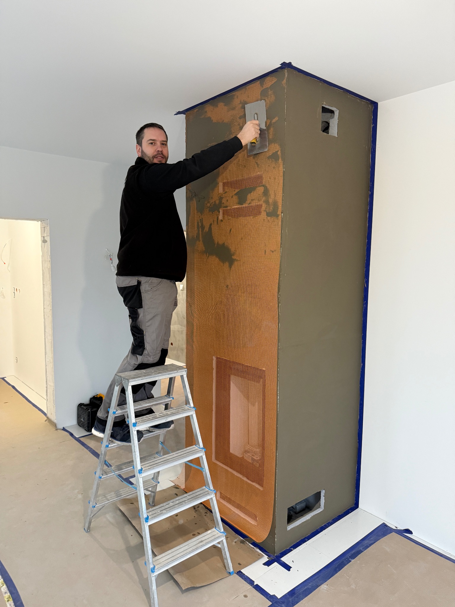 Mężczyzna na drabinie, zacierający szarą zaprawą obudowę kominka z kratką wzmacniającą, zabezpieczoną niebieską taśmą malarską.